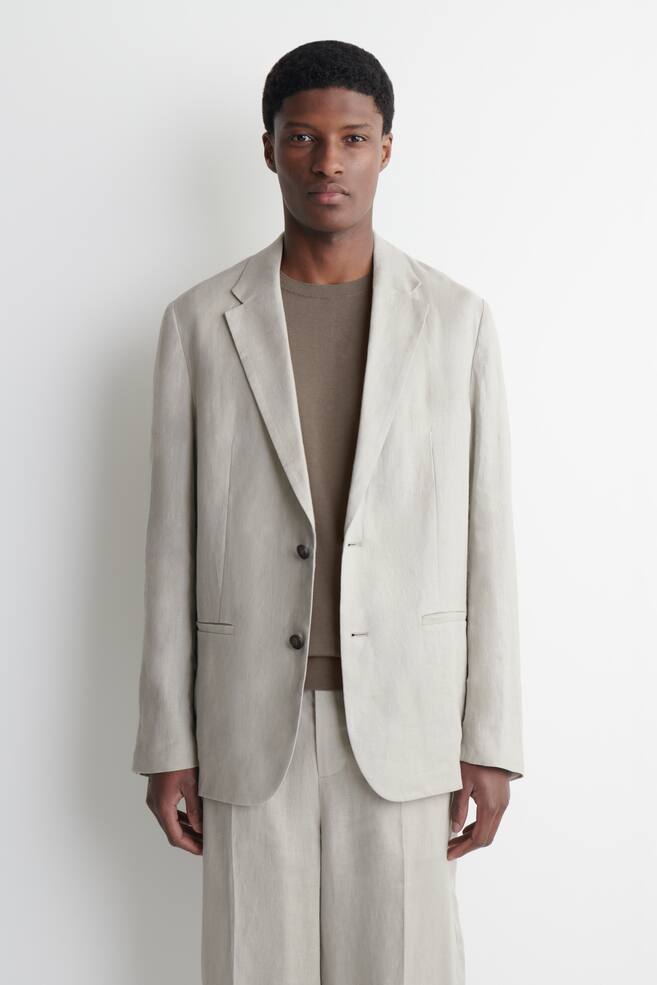 A light beige textured single-breasted blazer and matching wide-leg trousers are showcased, facing forward. The blazer features notch lapels, two dark buttons, and two flap pockets, and is worn over a brown crew neck t-shirt. - 1