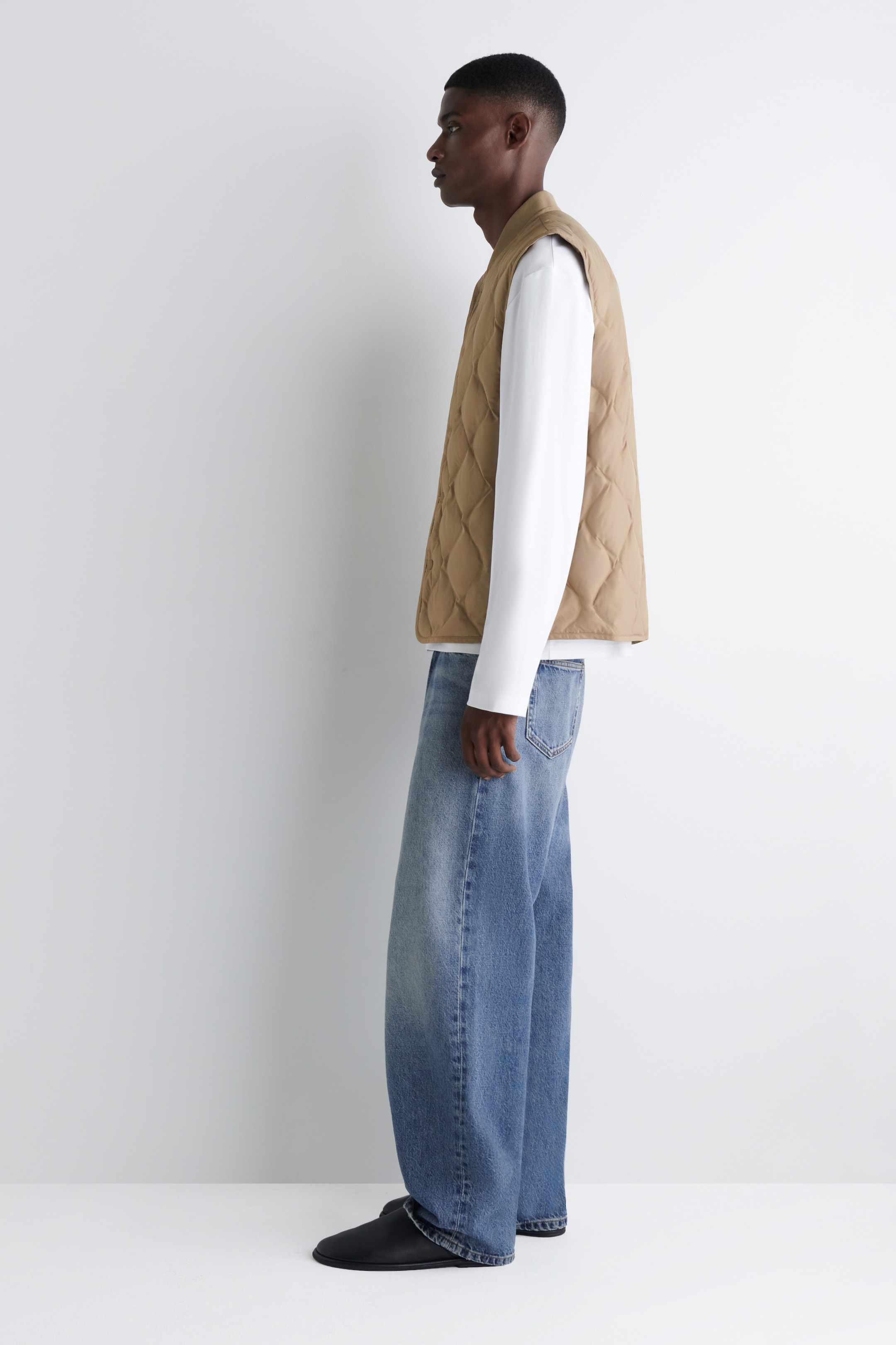 Agrandir l'image: Un gilet matelassé brun clair est présenté de profil, superposé à une chemise blanche à manches longues et associé à un jean délavé, ample et des chaussures plates noires.