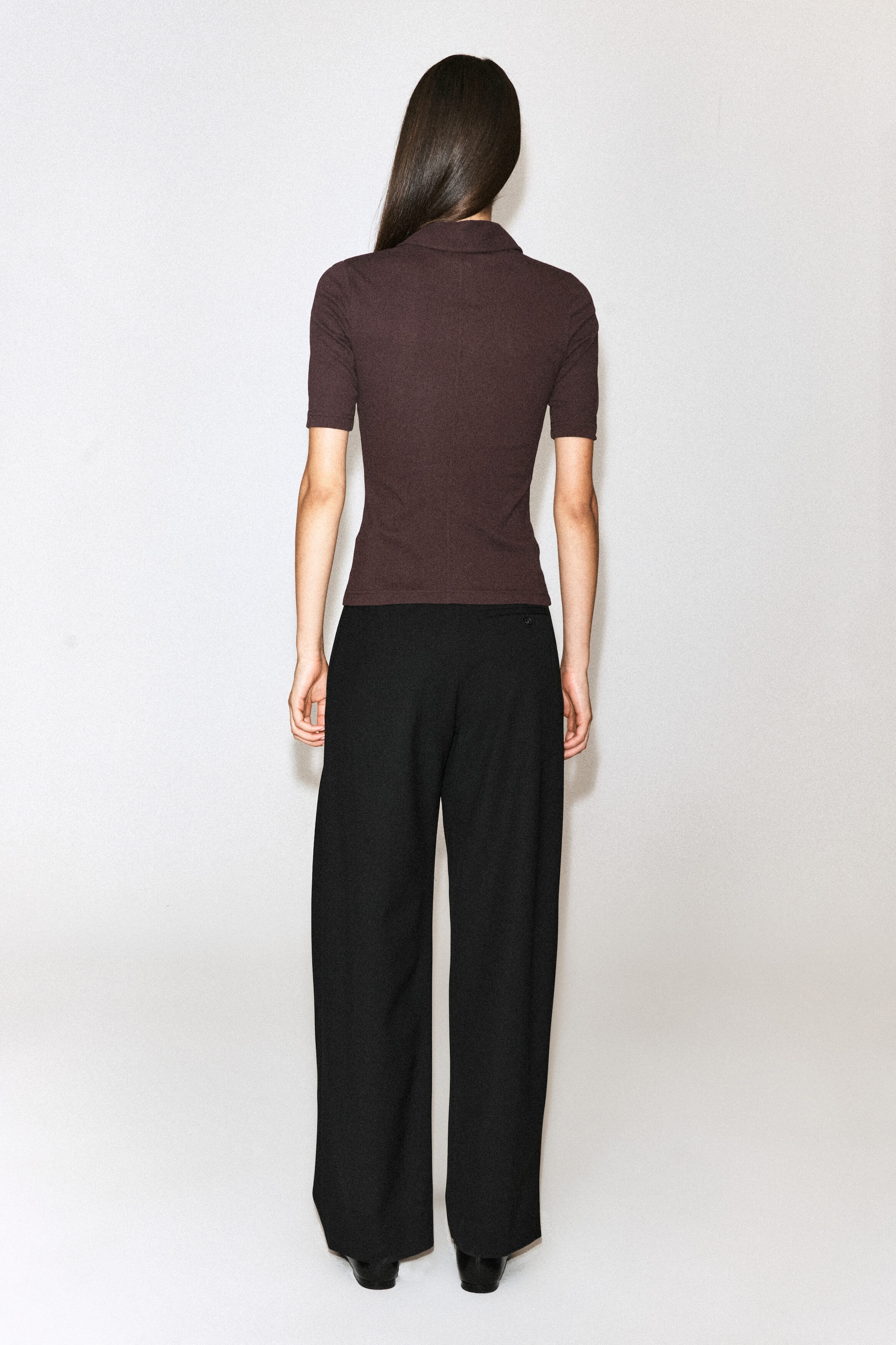 A dark brown, sculpted cotton jersey shirt with a fitted silhouette, short sleeves, and a collar is worn by the model, shown from the back. The shirt is styled with wide-leg black trousers. A dark brown, sculpted cotton jersey shirt with a fitted silhouette, short sleeves, and a collar is worn by the model, shown from the back. The shirt is styled with wide-leg black trousers.