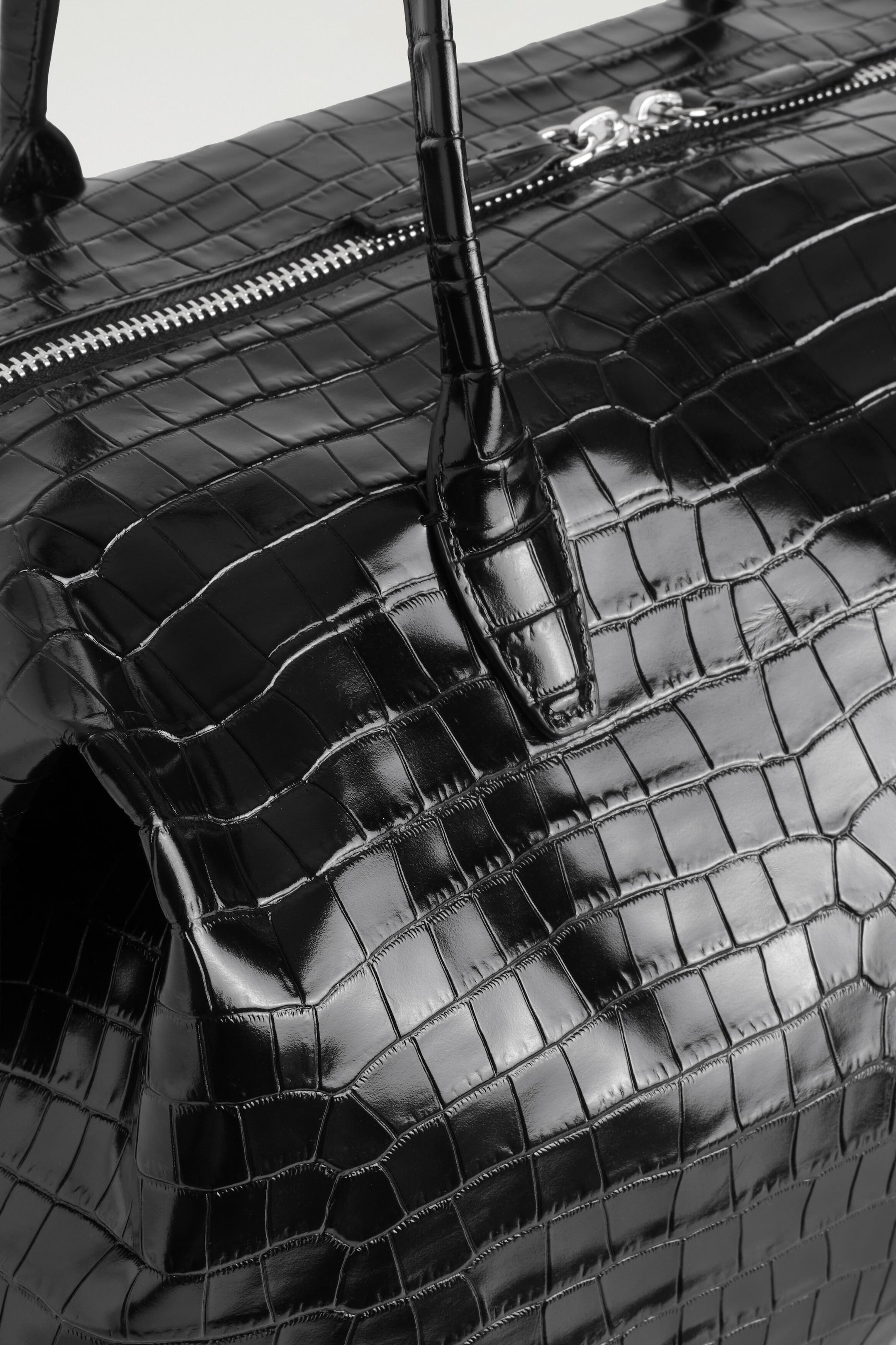 The Frieze Bowling Bag in black croc-effect leather is shown in an angled close-up view, highlighting its glossy, embossed texture. The bag features a silver-tone metal zip closure along the top and a matching rolled top handle. The Frieze Bowling Bag in black croc-effect leather is shown in an angled close-up view, highlighting its glossy, embossed texture. The bag features a silver-tone metal zip closure along the top and a matching rolled top handle.