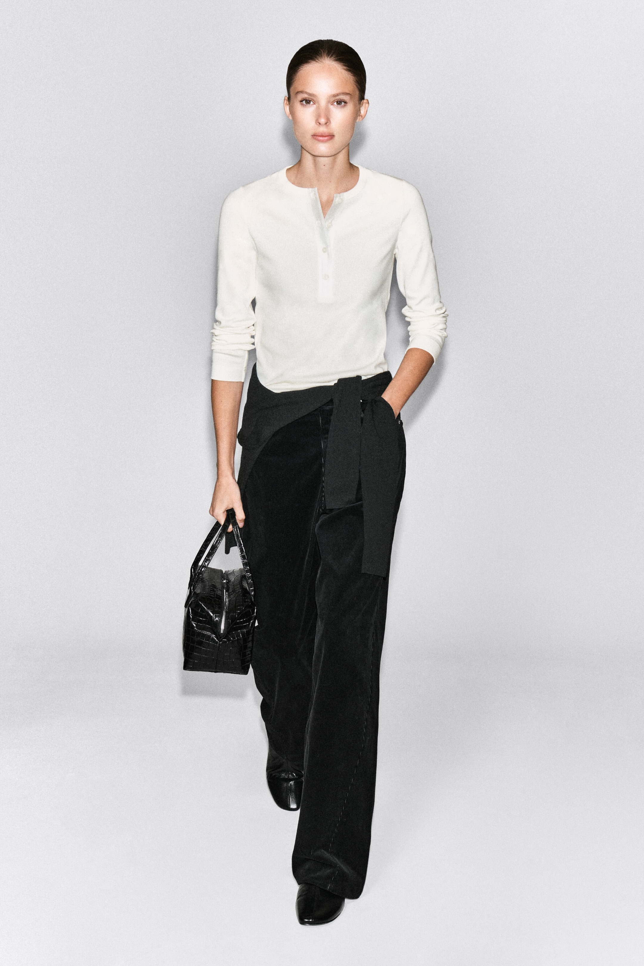 A ribbed cotton Henley top in off-white is worn by the model, featuring long sleeves, a crew neck, and a button placket. The top is styled with black wide-leg trousers, a black sweater tied around the waist, and a black textured handbag carried by hand. A ribbed cotton Henley top in off-white is worn by the model, featuring long sleeves, a crew neck, and a button placket. The top is styled with black wide-leg trousers, a black sweater tied around the waist, and a black textured handbag carried by hand.