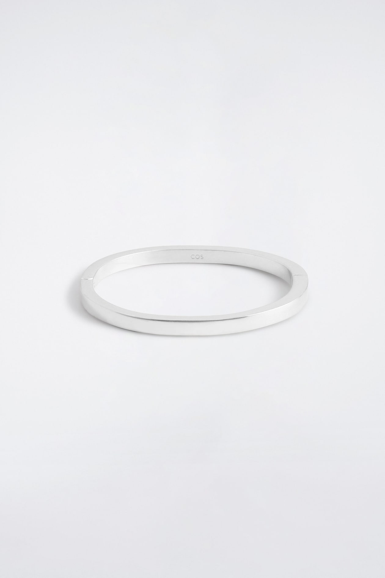 Un bracelet rigide minimaliste aux reflets argentés est posé à plat, vu de face, mettant en valeur son profil net à bords plats et sa finition métallique brossée. De discrètes charnières sont visibles sur les côtés, et un petit logo est gravé à l’intérieur du bracelet.