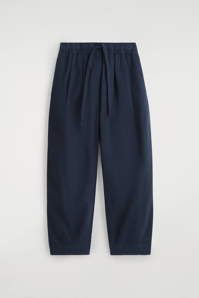 Marineblaue Hosen werden VORNE angezeigt, mit einer entspannten Passform und einer verkürzten Länge. Sie sind mit einem elastischen Kordelzugbund, Faltendetails unterhalb der Taille und entspannten Beinen, die sich zu den Knöcheln hin verjüngen, gestaltet. - 1