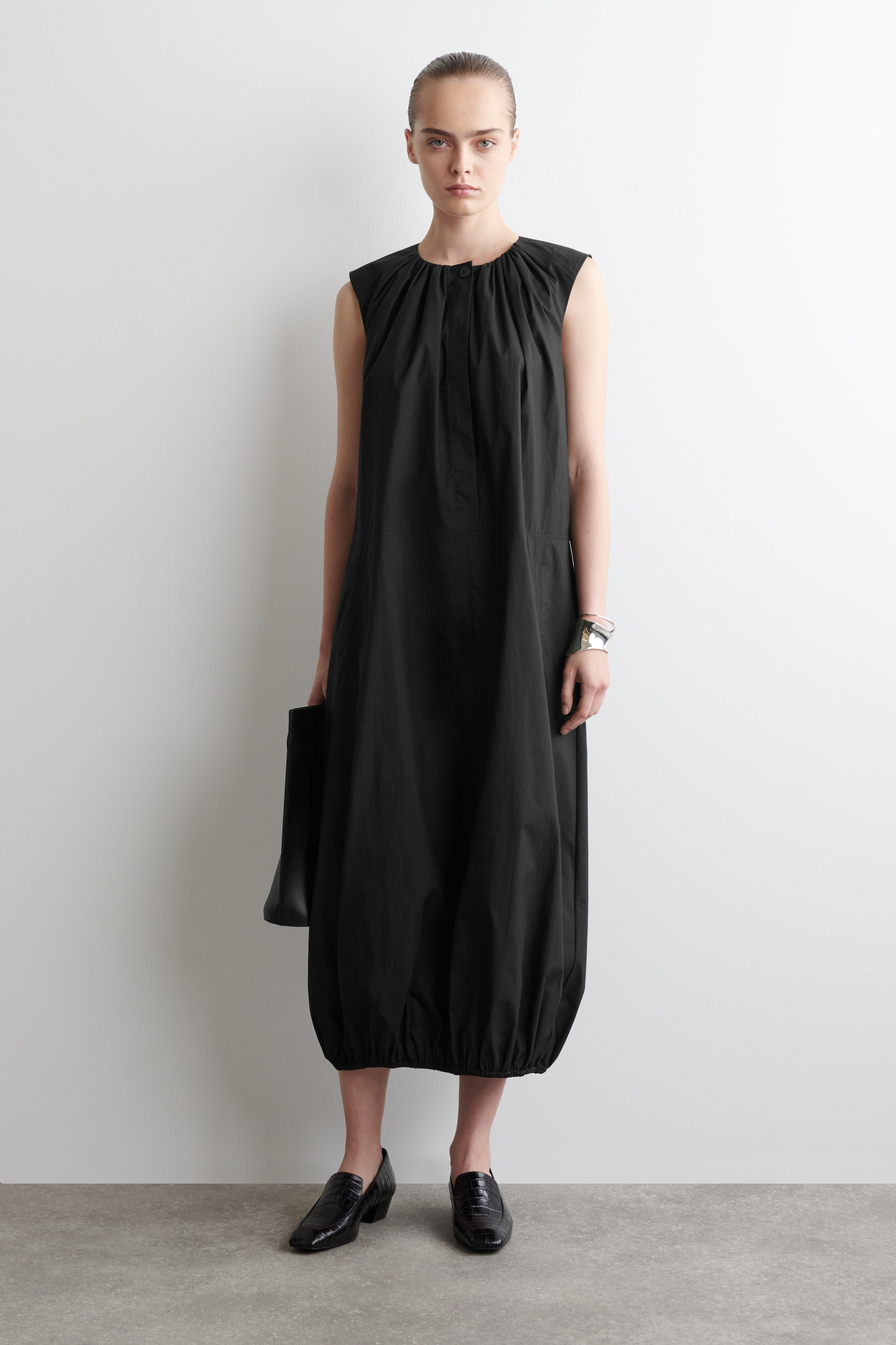 Agrandir l'image: Une robe midi noire est présentée de FACE, avec une coupe décontractée, un design sans manches et un col rond froncé avec une fermeture à bouton visible au centre avant. Elle est conçue avec des poches latérales discrètes et un ourlet bouillonné froncé, créant une silhouette pleine et arrondie.