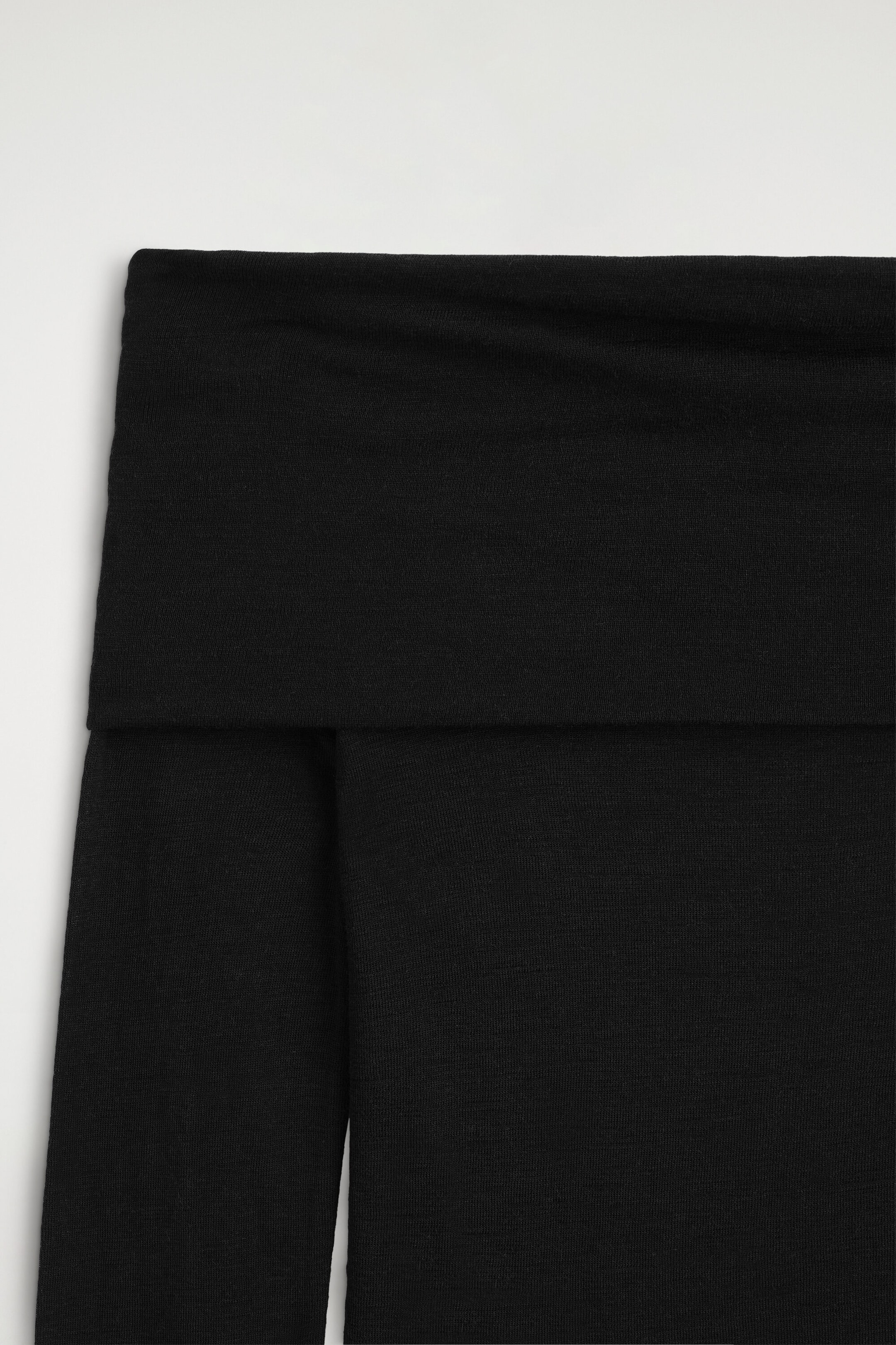 A close-up front view of the black off-the-shoulder merino wool top highlights its wide, folded-over neckline and fine-knit texture. The garment features a seamless, double-layered band that drapes over the upper bodice and sleeves.