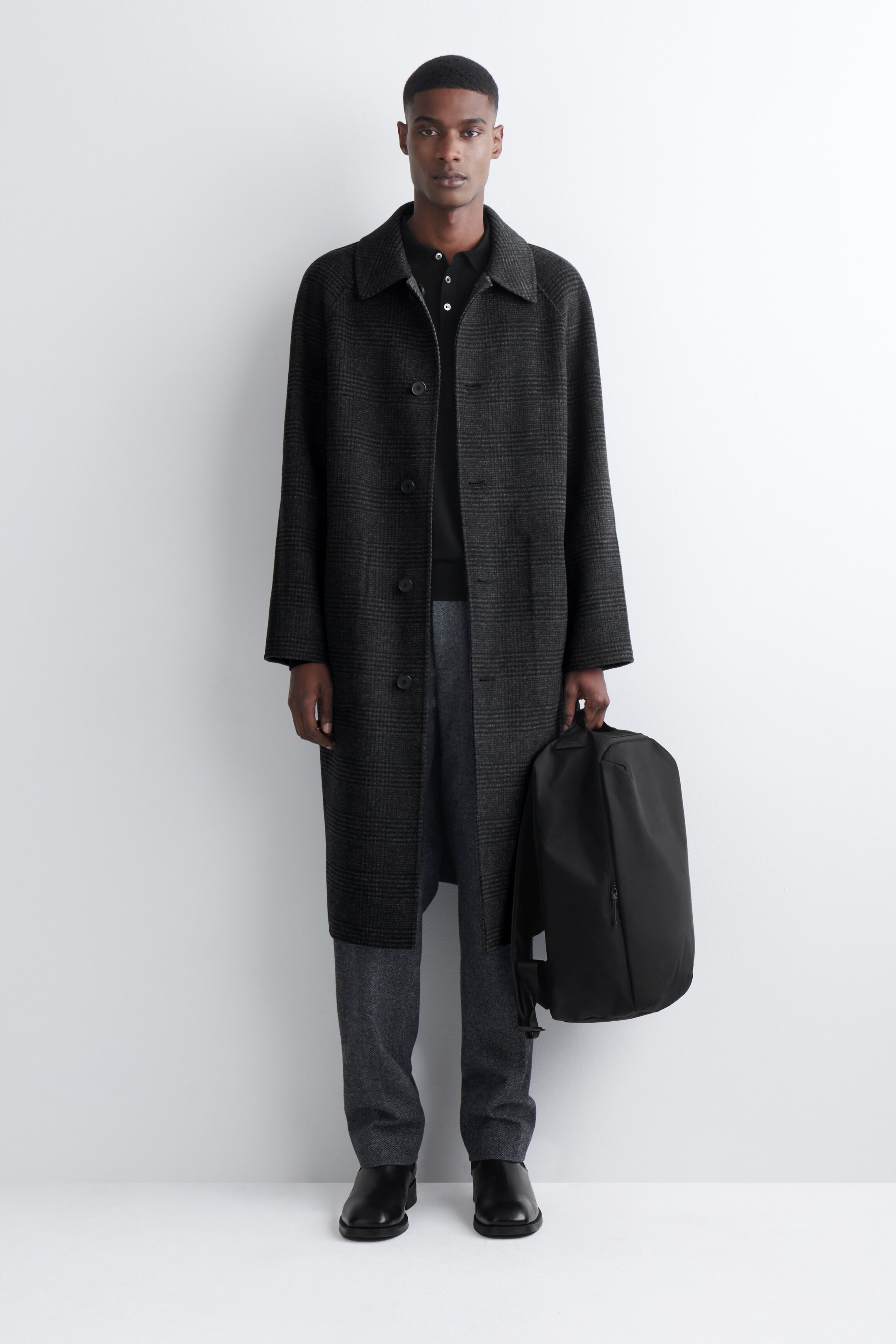 Agrandir l'image: Un manteau à carreaux gris foncé est présenté de face, avec un col évasé, une fermeture à boutons sur le devant et des manches longues. Il offre une coupe décontractée, s'étendant juste en dessous du genou, et est superposé à un polo foncé et un pantalon gris.