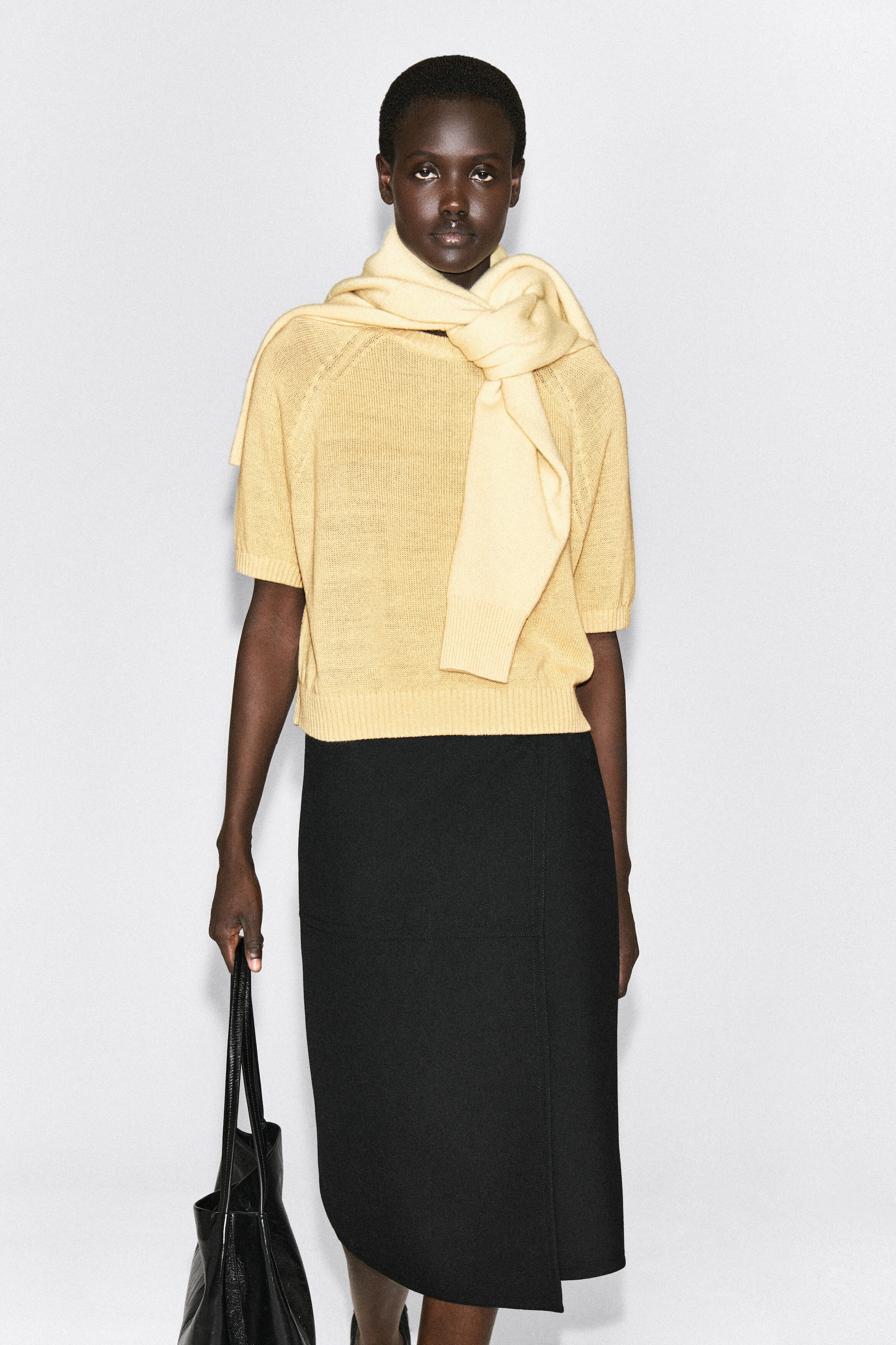 The shrunken knitted linen t-shirt is shown from the front, highlighting its light yellow, semi-sheer knit structure with short raglan sleeves, ribbed cuffs, and a ribbed hem. A cream-coloured knit sweater is draped over the shoulders and knotted at the neck, layering over the t-shirt’s neckline. The shrunken knitted linen t-shirt is shown from the front, highlighting its light yellow, semi-sheer knit structure with short raglan sleeves, ribbed cuffs, and a ribbed hem. A cream-coloured knit sweater is draped over the shoulders and knotted at the neck, layering over the t-shirt’s neckline.