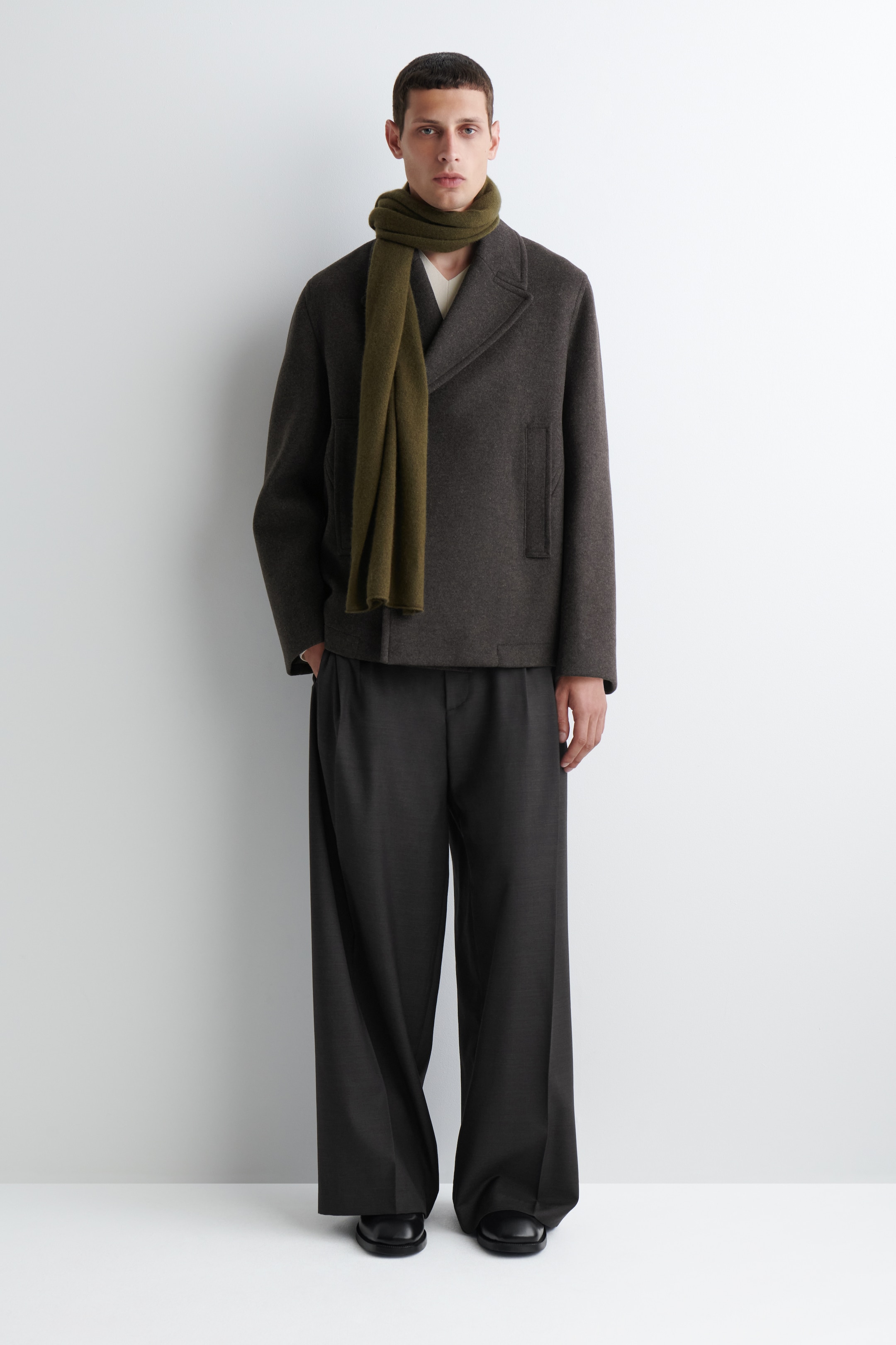 Größeres Bild anzeigen: Das Produkt ist ein komplettes Outfit, das nach vorne gerichtet ist und aus einem dunkelgrauen, kurzen, zweireihigen Sakko mit breiten Kerbrevers und seitlichen Pattentaschen besteht, das über einem hellen Unterhemd getragen wird. Ein olivgrüner Schal ist um den Hals drapiert und ergänzt die dunkelgrauen, weit geschnittenen, plissierten Hosen und schwarzen, schlichten Schuhe.