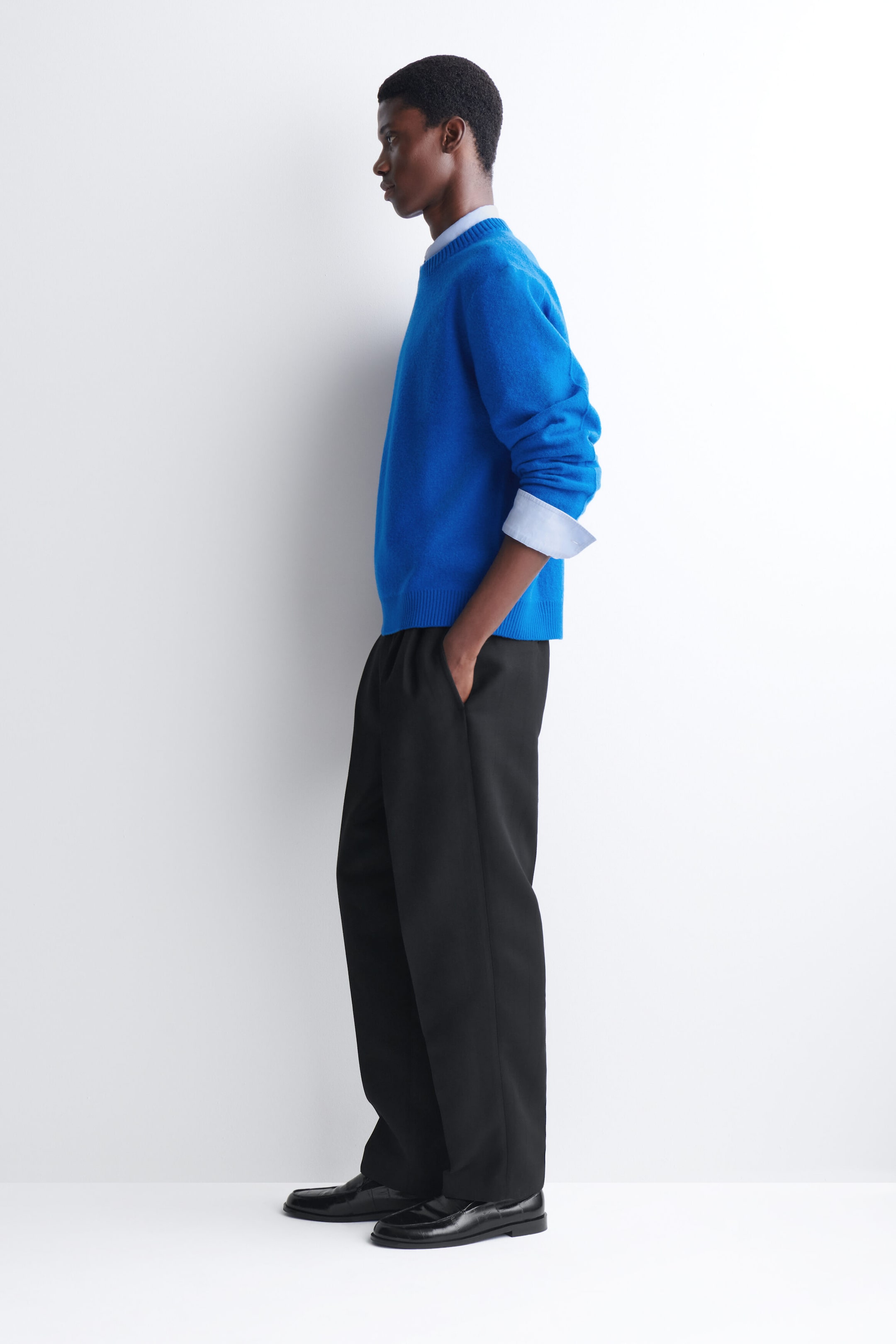 A bright blue, relaxed-fit crewneck sweater is being worn by the model, who is standing in profile. The knitwear is layered over a light blue collared shirt with folded cuffs and paired with black trousers. A bright blue, relaxed-fit crewneck sweater is being worn by the model, who is standing in profile. The knitwear is layered over a light blue collared shirt with folded cuffs and paired with black trousers.