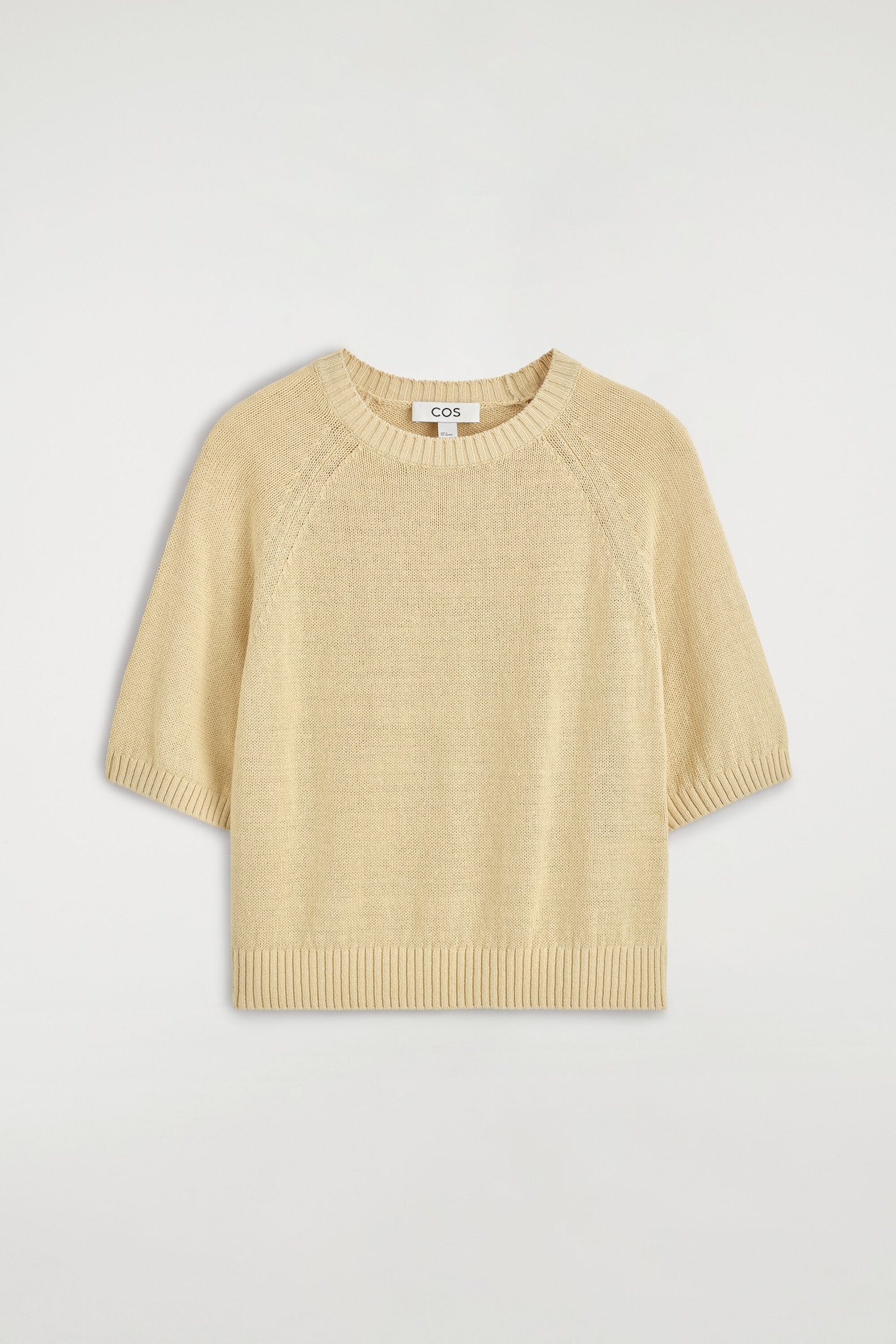 A shrunken knitted linen T-shirt in a pale beige colour is facing forward, showcasing its fine-knit texture, ribbed crew neck, and short raglan sleeves with ribbed cuffs. It features a matching ribbed hem that defines its shrunken silhouette.