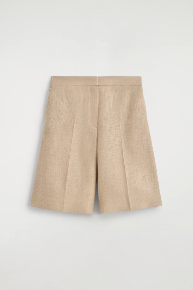 A pair of light beige shorts is presented facing front, featuring a longline silhouette with press-creased straight legs. The shorts display a flat waistband and a subtly textured fabric. - 1