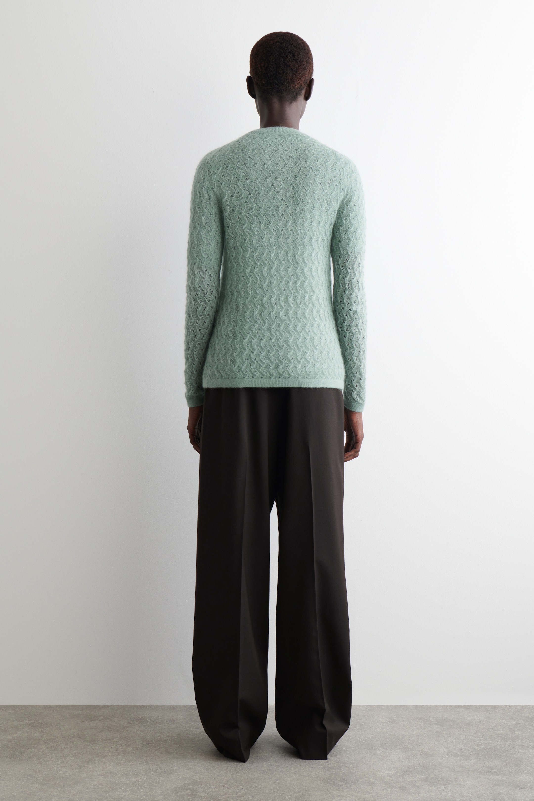 Grösseres Bild ansehen: Ein hellgrüner Rundhalspullover wird von hinten gezeigt, mit einem strukturierten Strickmuster und einem weichen, flauschigen Finish. Er wird mit weit geschnittenen, dunkelbraunen Hosen gestylt.