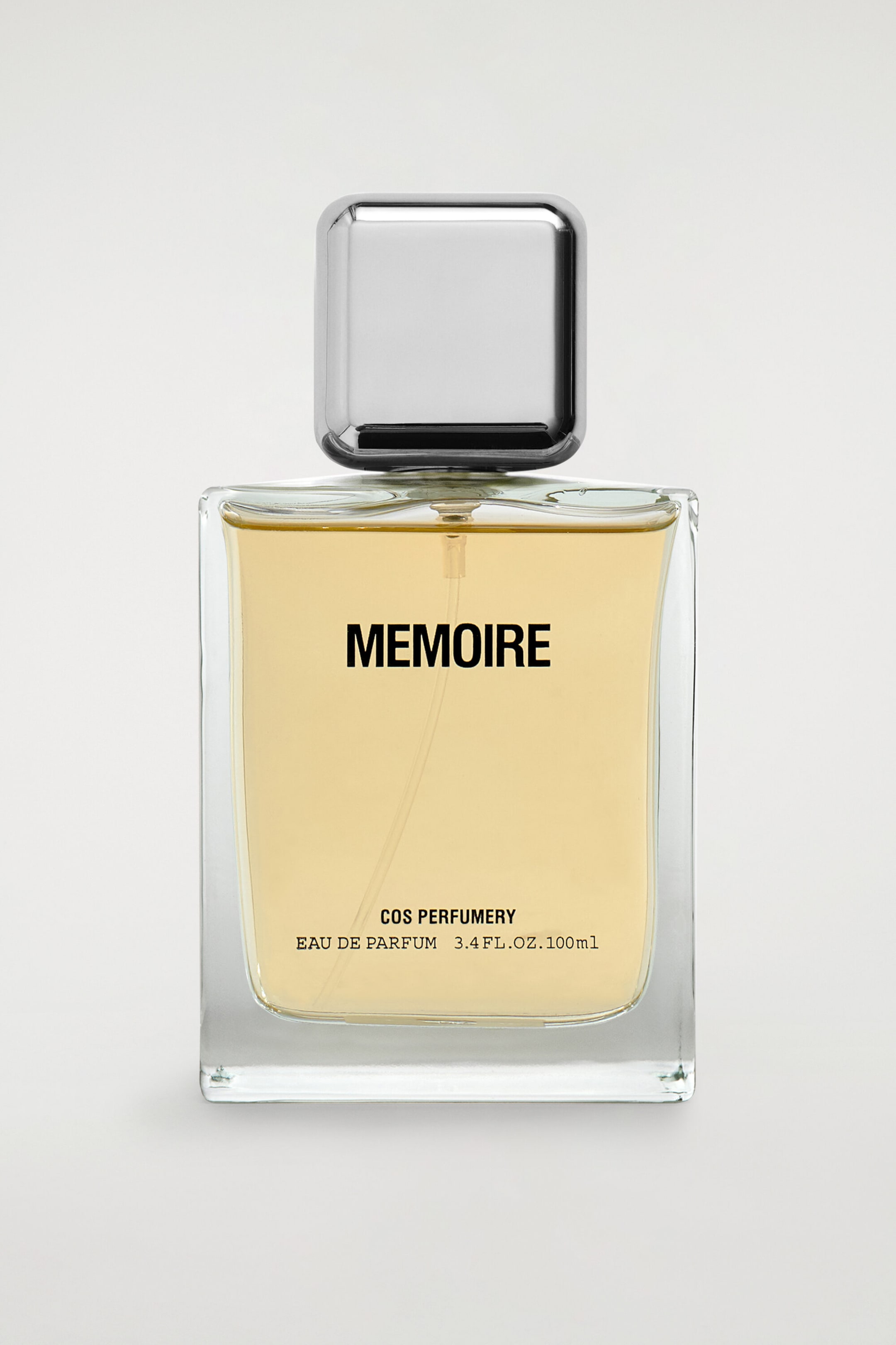 The COS Perfumery Eau de Parfum, 100ml, is standing front-facing, featuring a rectangular clear glass bottle filled with a translucent, light amber liquid and topped with a square, polished silver-toned cap. The front of the bottle displays the printed name "MEMOIRE" in bold black letters, with "COS PERFUMERY" and "EAU DE PARFUM 3.4 FL. OZ. 100ml" positioned near the bottom.