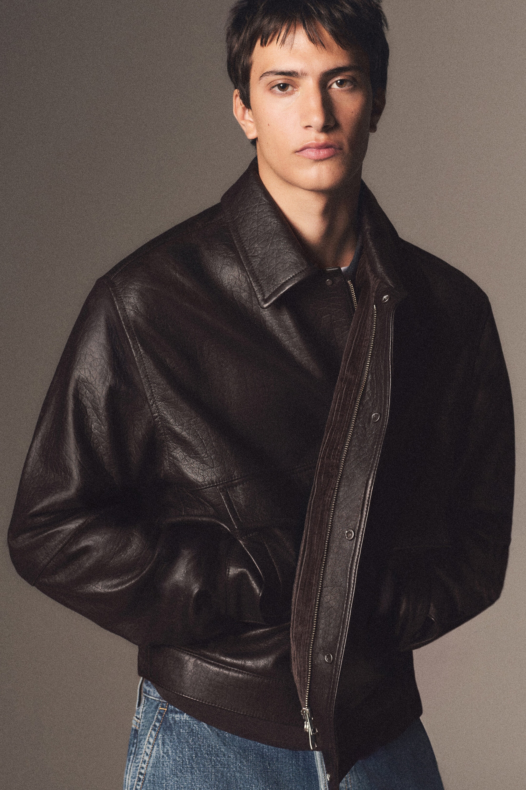 A dark brown leather flight jacket is shown from the front, featuring a textured finish, a classic point collar, and a front zip closure with an overlapping snap-button placket. The jacket features a relaxed silhouette with a ribbed hem and side welt pockets, and is styled partially open over blue jeans. A dark brown leather flight jacket is shown from the front, featuring a textured finish, a classic point collar, and a front zip closure with an overlapping snap-button placket. The jacket features a relaxed silhouette with a ribbed hem and side welt pockets, and is styled partially open over blue jeans.