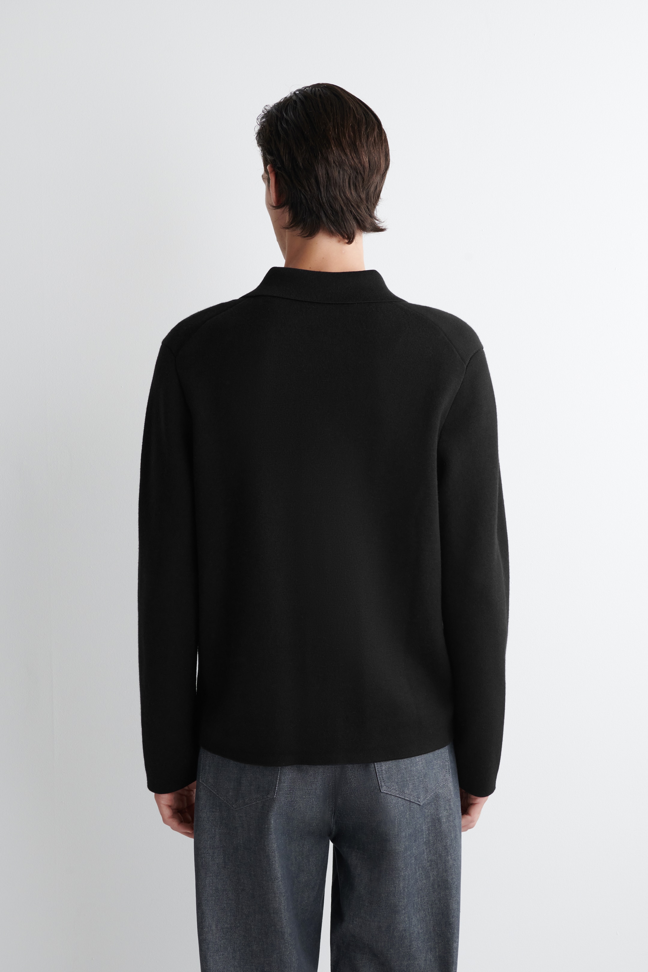 Agrandir l'image: Un polo en tricot noir à manches longues est montré de dos, avec un col polo classique et des manches longues lisses.