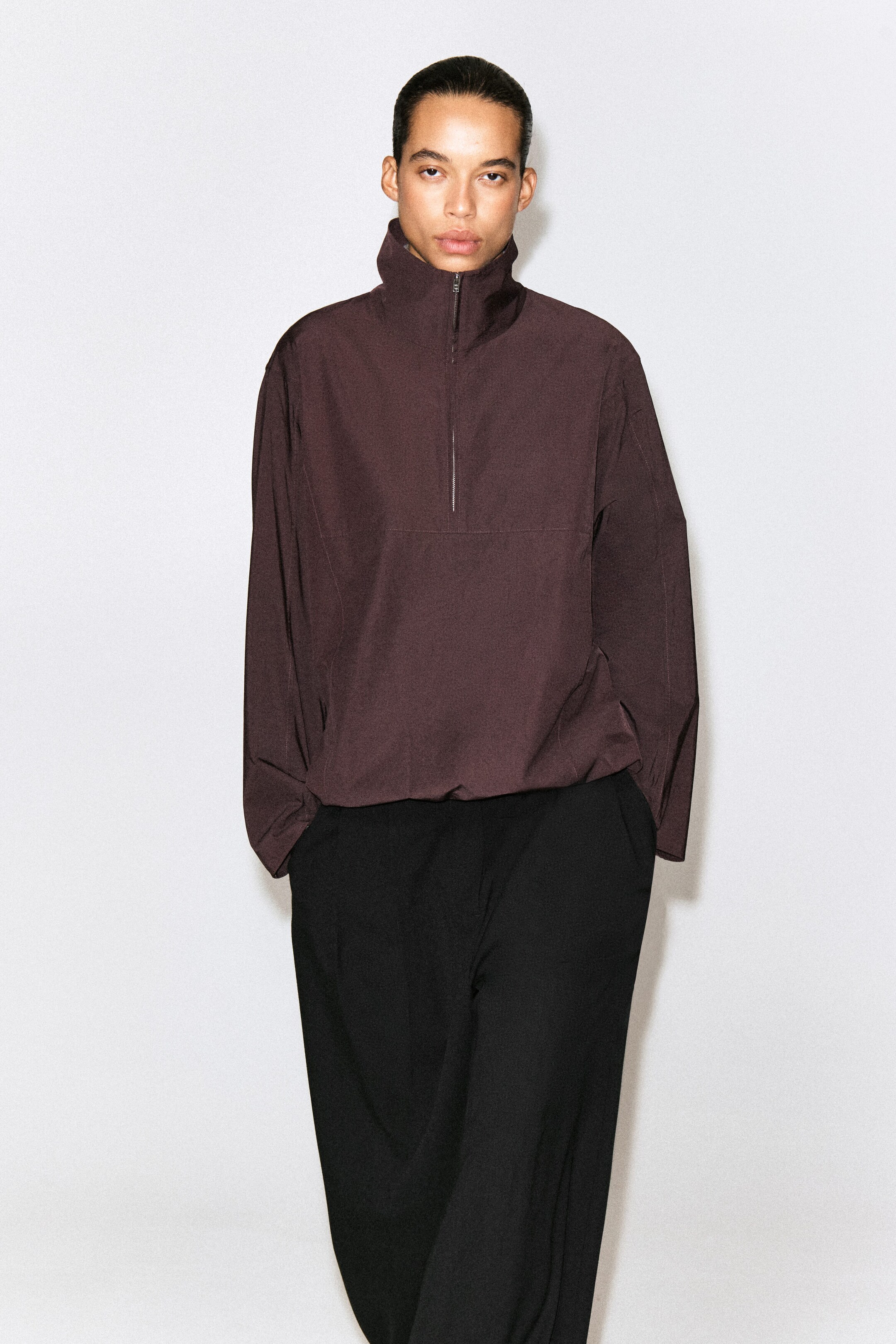 A dark plum half-zip pullover shirt is shown from the front, featuring a high stand collar with a silver-tone zipper, long sleeves, and a gathered drawstring hem. A dark plum half-zip pullover shirt is shown from the front, featuring a high stand collar with a silver-tone zipper, long sleeves, and a gathered drawstring hem.