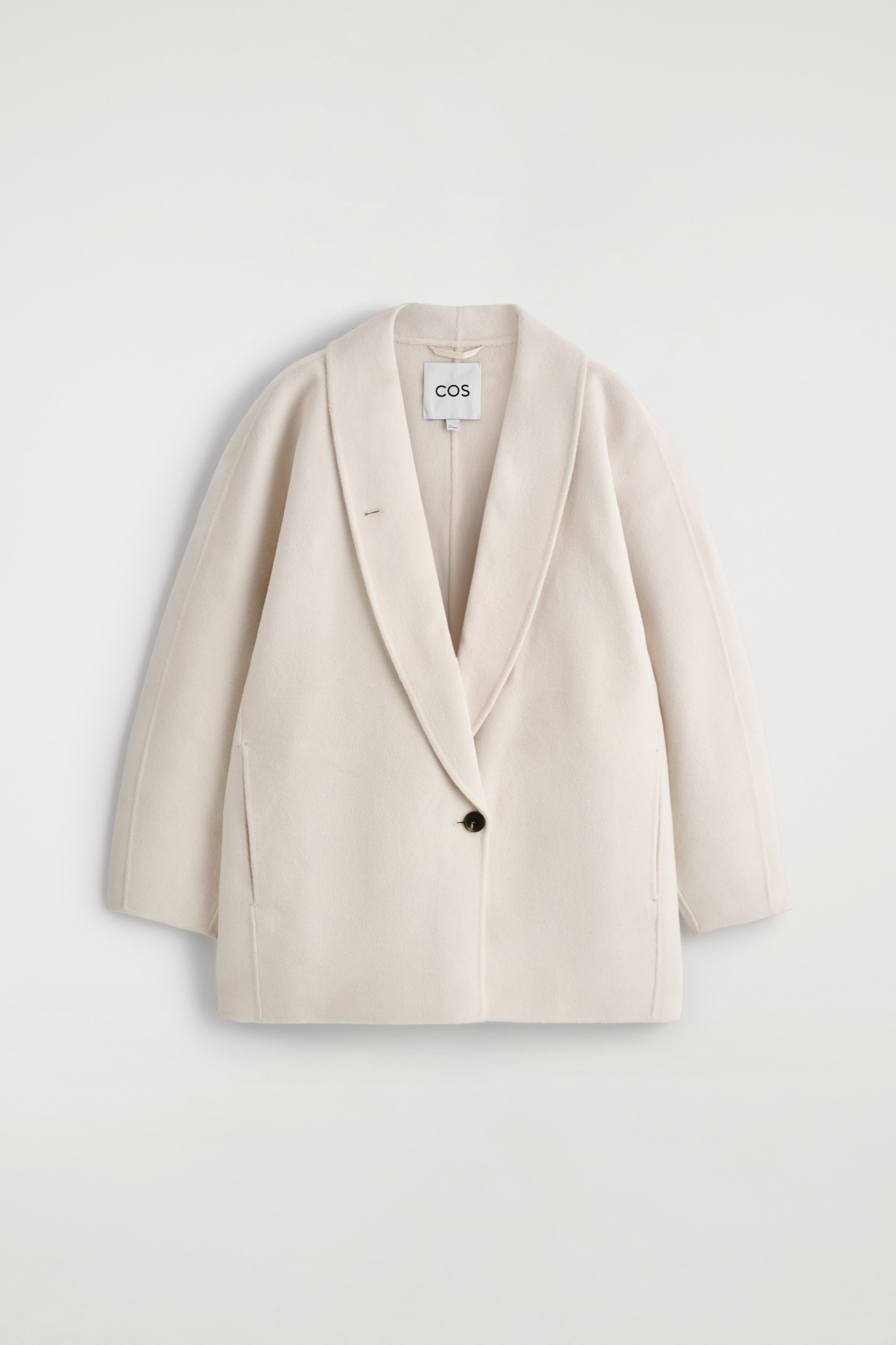 Größeres Bild ansehen: Eine hellcremefarbene Cabanjacke wird VON VORNE gezeigt, mit einem breiten Schalkragen und einem einzelnen dunklen Knopfverschluss. Sie zeigt eine entspannte Passform mit schrägen Seitentaschen und reicht bis zur Hüfte.