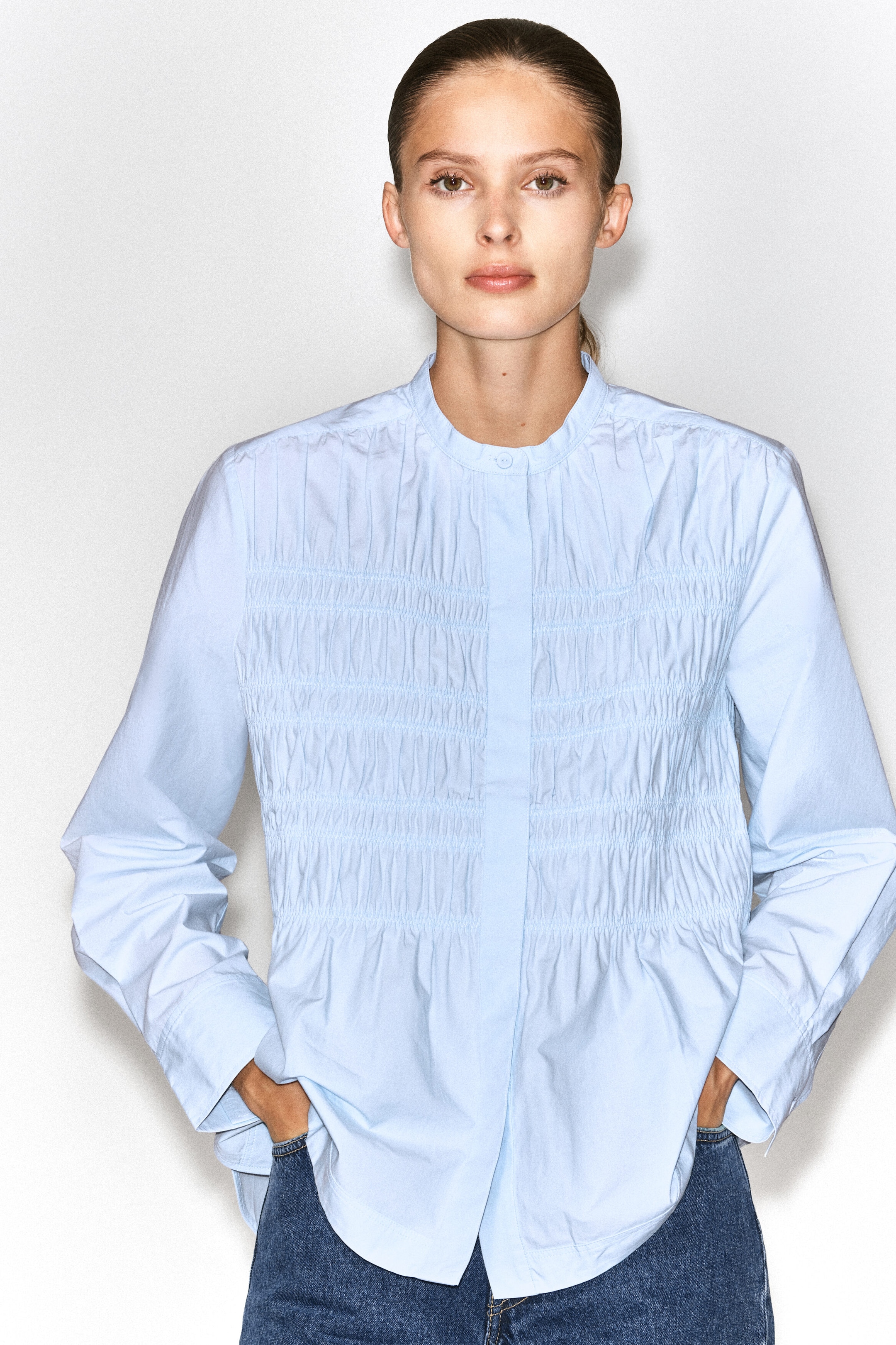 A light blue ruched cotton blouse is being worn by the model, who is posing with her hands in the pockets of her blue jeans. The blouse features a band collar, long sleeves, and vertical smocked detailing across the front chest. A light blue ruched cotton blouse is being worn by the model, who is posing with her hands in the pockets of her blue jeans. The blouse features a band collar, long sleeves, and vertical smocked detailing across the front chest.