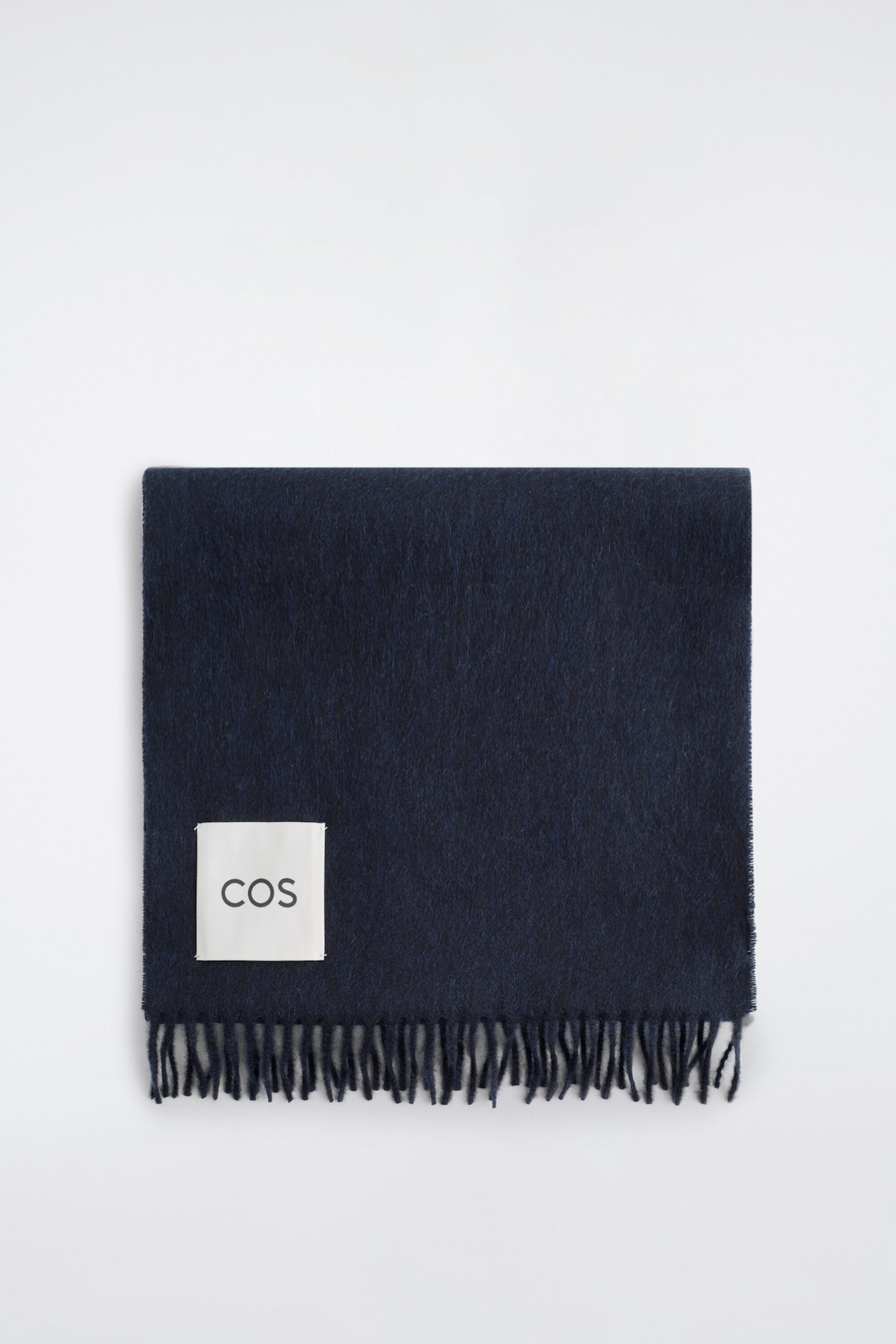 A dark navy-blue fringed cashmere scarf is shown folded flat from the front, highlighting its soft, brushed texture and twisted fringe detailing along the bottom edge. A square white brand label featuring the text "COS" is stitched onto the lower-left corner.