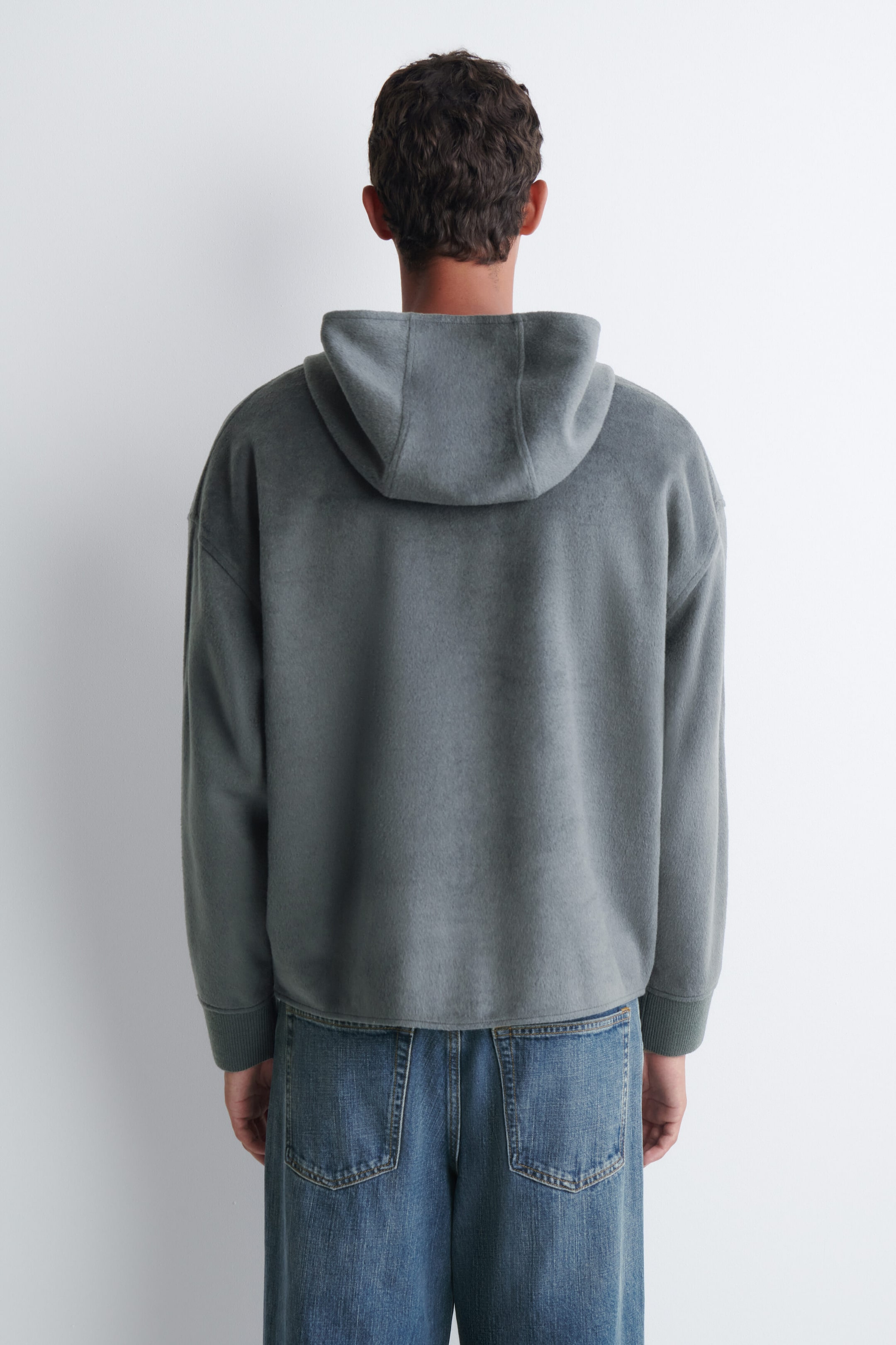 Un chandail à capuchon en molleton gris-vert est porté par le mannequin, vu de dos. Le chandail présente une coupe décontractée, des épaules tombantes, des poignets côtelés et une capuche, et il est agencé avec un jean en denim bleu. Un chandail à capuchon en molleton gris-vert est porté par le mannequin, vu de dos. Le chandail présente une coupe décontractée, des épaules tombantes, des poignets côtelés et une capuche, et il est agencé avec un jean en denim bleu.