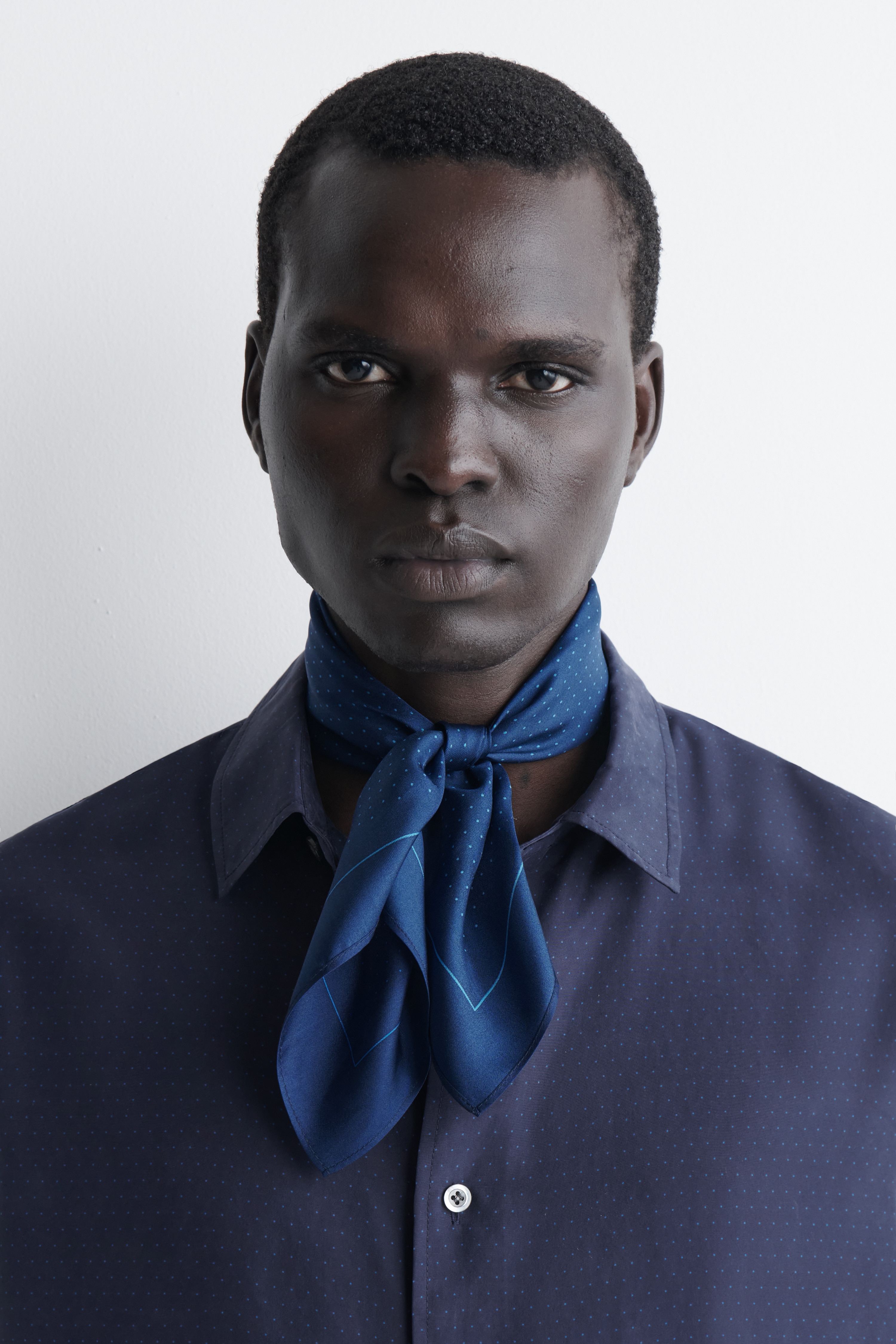 A dark blue silk pocket square lies flat, facing forwards, with one corner folded back to show the reverse side. The pocket square features a pattern of tiny light blue polka dots framed by a matching thin blue inner border, and finished with stitched edges.