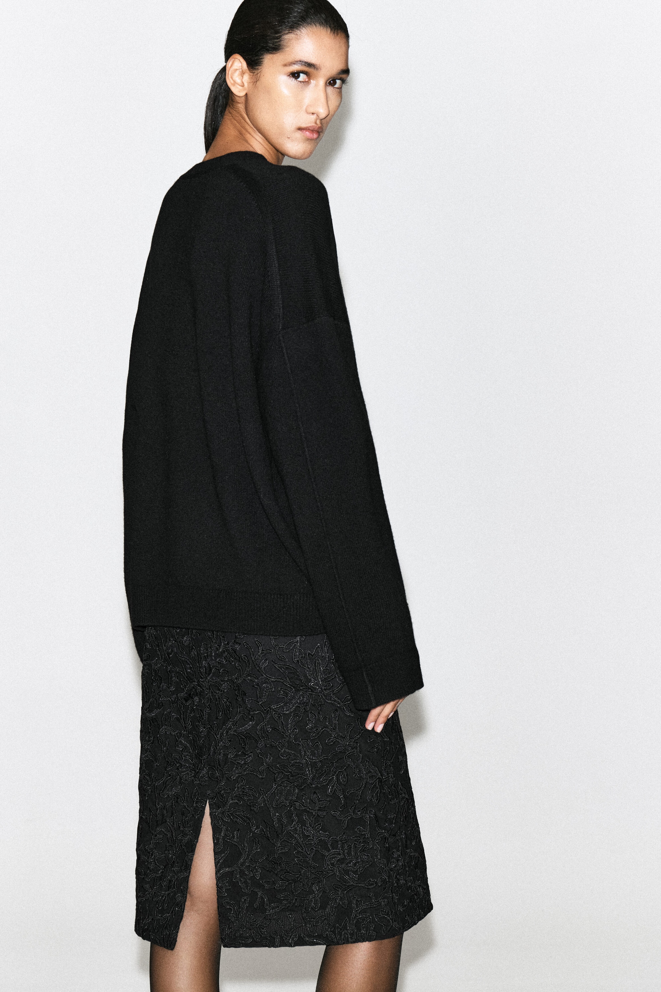 Un cardigan nero in maglia cade morbidamente sul retro, mettendo in risalto la sua vestibilità rilassata, le spalle scese e il bordo a coste. Il cardigan è abbinato a una gonna midi nera dalla texture materica, caratterizzata da uno spacco posteriore.