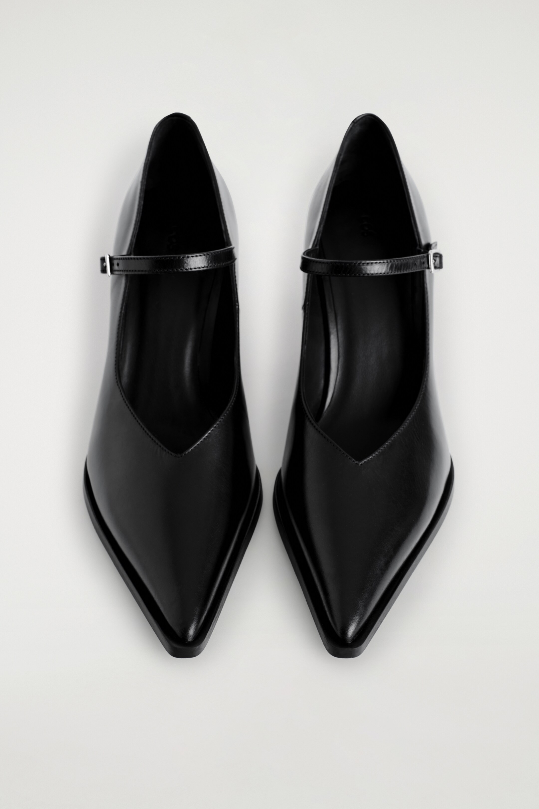 Agrandir l'image: Chaussures Mary Jane à talons en cuir. Une paire de chaussures Mary Jane à talons en cuir noir est présentée en vue de dessus, avec des bouts pointus, un décolleté en V profond et une fine bride sur le cou-de-pied avec une boucle argentée.