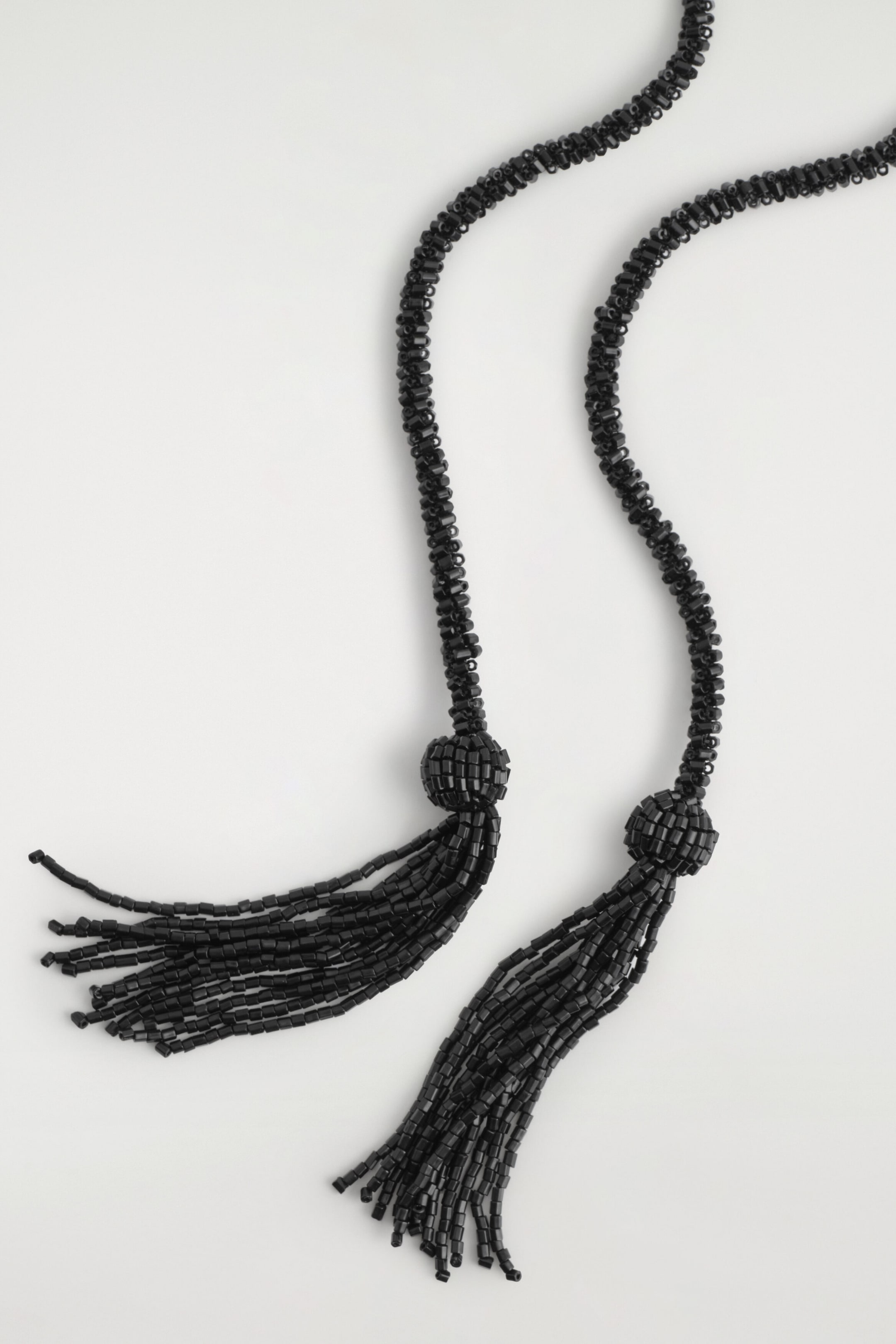 A black beaded necklace is shown lying flat, featuring long strands made of small, glossy black beads. The strands end in decorative, bead-wrapped knots with multiple fine beaded tassels hanging down.