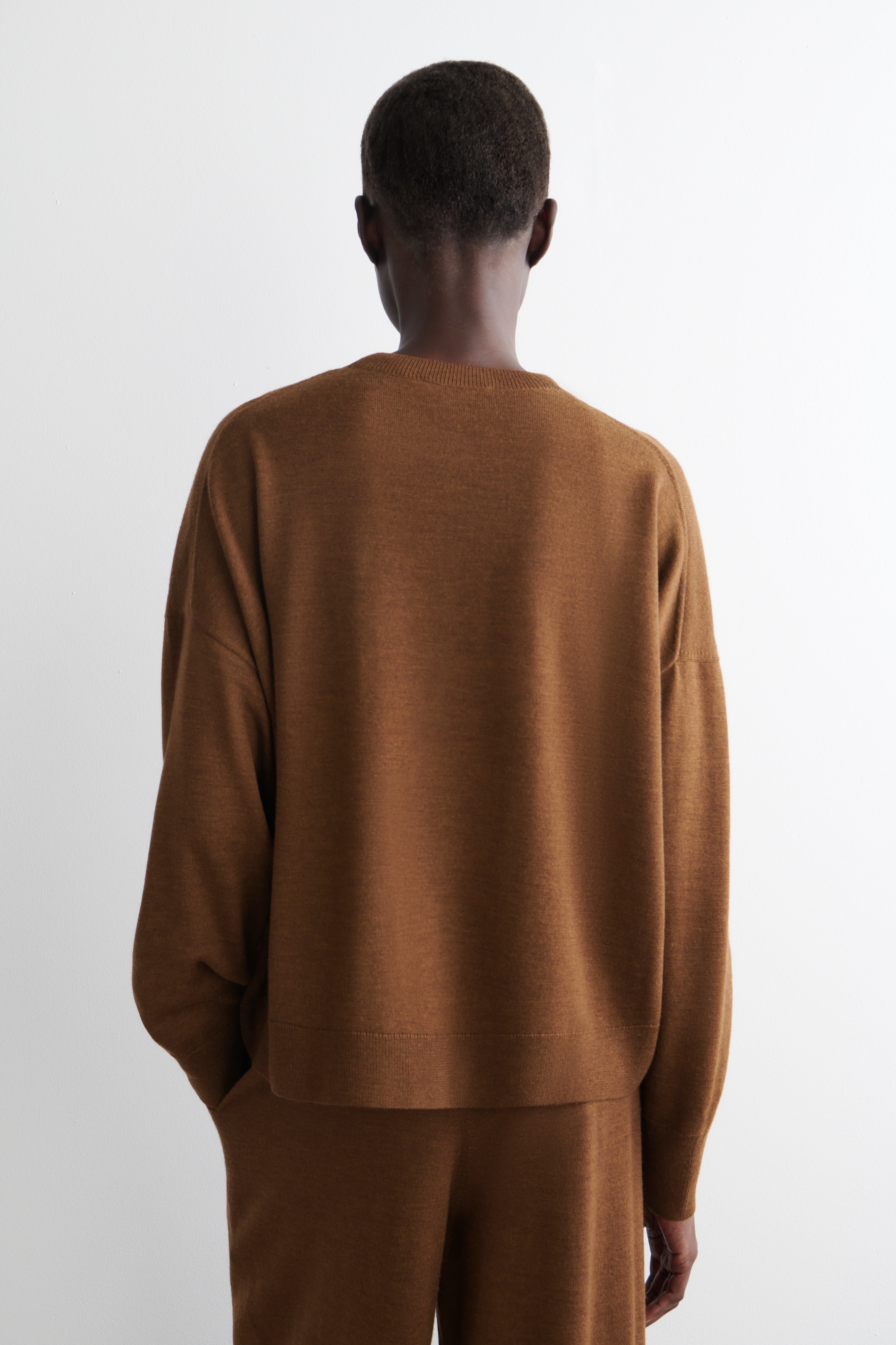 Agrandir l'image: Un pull en tricot marron et un pantalon assorti sont vus de dos. Le pull présente une coupe décontractée avec un col rond, des manches longues et un ourlet côtelé. Le pantalon coordonné montre également une coupe décontractée, avec une main reposant dans une poche latérale.