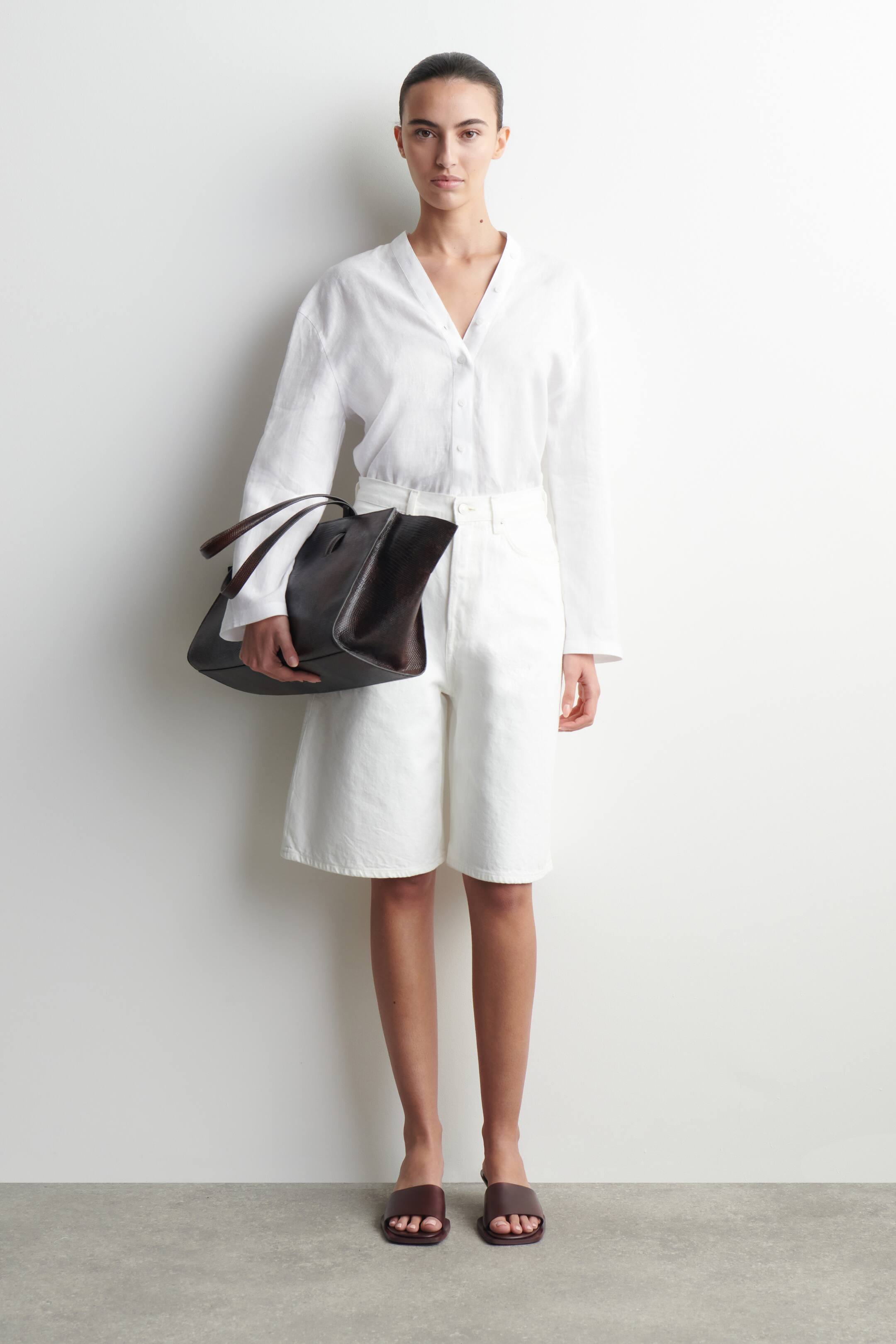 Visa större bild: En mörkbrun handväska med reptilmönster hålls upp, dess mjuka, ostrukturerade form visar upp två matchande topphandtag. Den är vinklad något åt sidan, vilket framhäver dess texturerade yta.