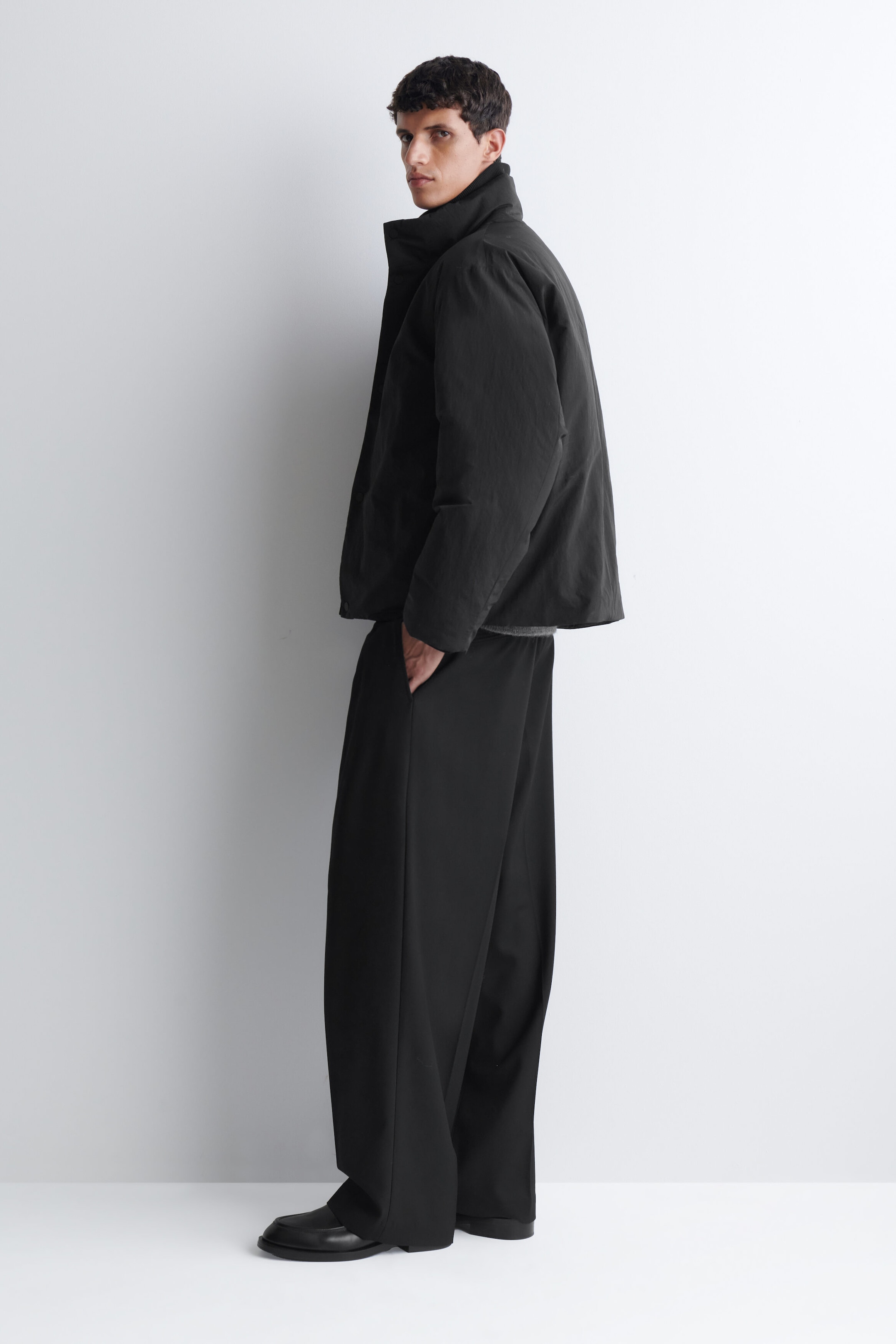A black padded funnel-neck down jacket is worn by the model, featuring a high, structured collar and a clean, minimalist design. The jacket is styled with wide-leg black trousers and black leather shoes, with the model standing in profile. A black padded funnel-neck down jacket is worn by the model, featuring a high, structured collar and a clean, minimalist design. The jacket is styled with wide-leg black trousers and black leather shoes, with the model standing in profile.