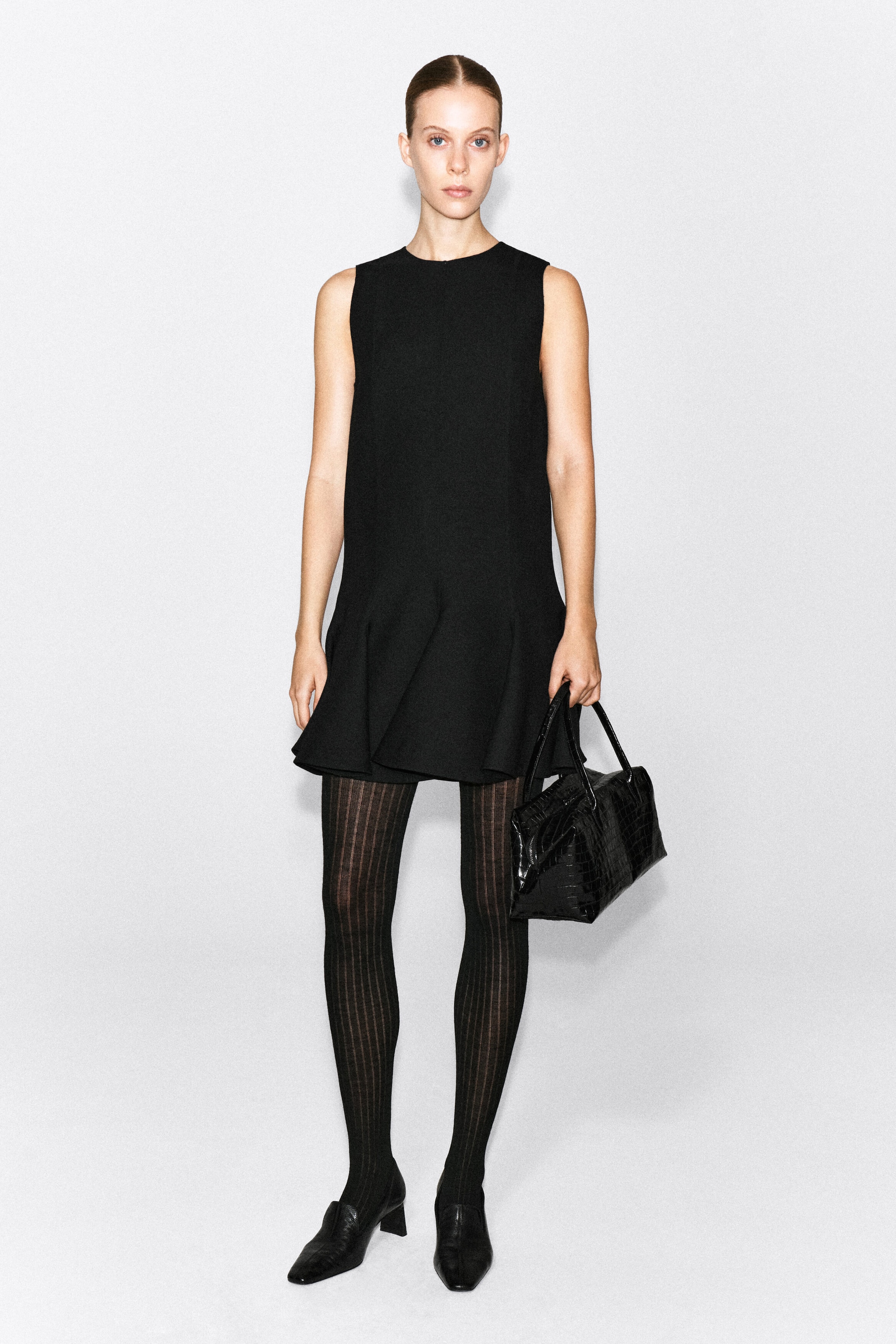 A black sleeveless mini dress is shown from the front, featuring a high crew neck, a structured shift silhouette, and a flared, dropped peplum hem. The dress is styled with sheer black ribbed tights and black heeled loafers. A black sleeveless mini dress is shown from the front, featuring a high crew neck, a structured shift silhouette, and a flared, dropped peplum hem. The dress is styled with sheer black ribbed tights and black heeled loafers.