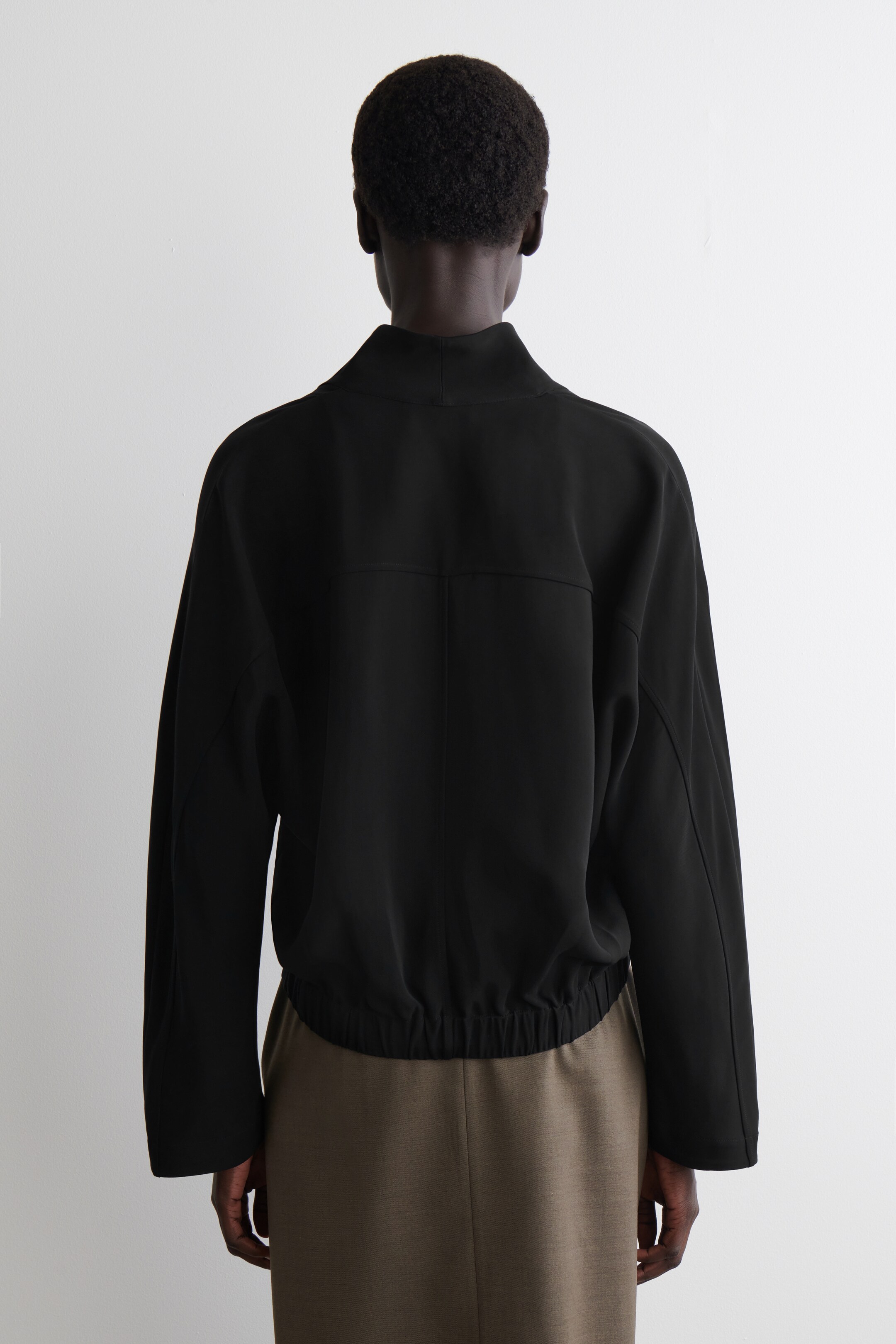 Agrandir l'image: Une veste noire est présentée de dos, avec un col montant, des épaules tombantes et une couture horizontale traversant le haut du dos. La veste est resserrée à la taille par un ourlet élastiqué et est portée sur un pantalon brun clair.