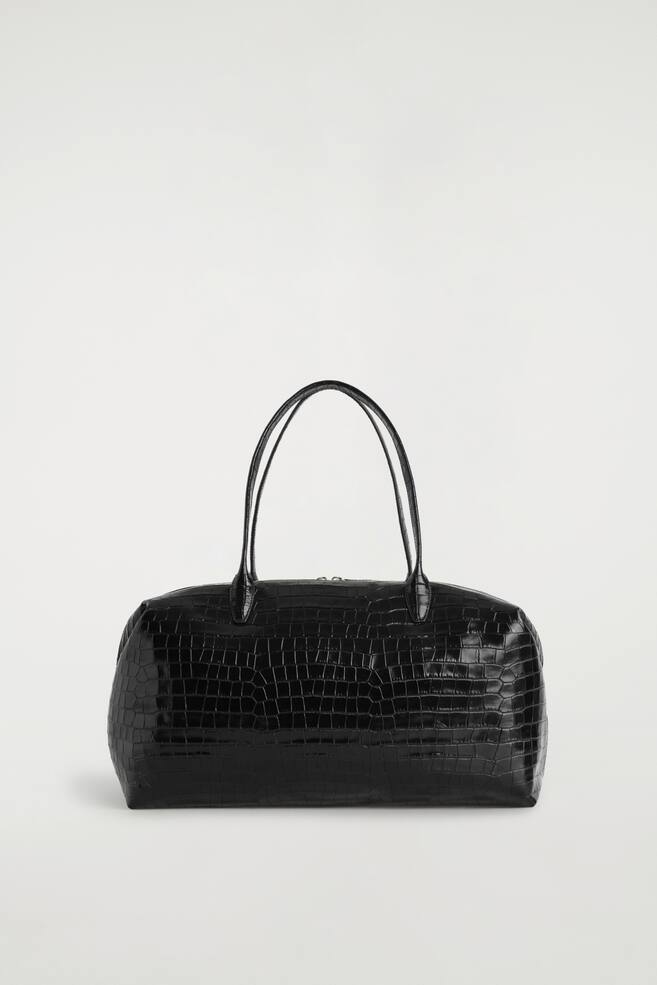The Frieze bowling bag in black croc-effect leather is shown from the front, showcasing its structured rectangular silhouette and glossy embossed texture. Two slim arched top handles extend upwards from the main body of the bag. - 1