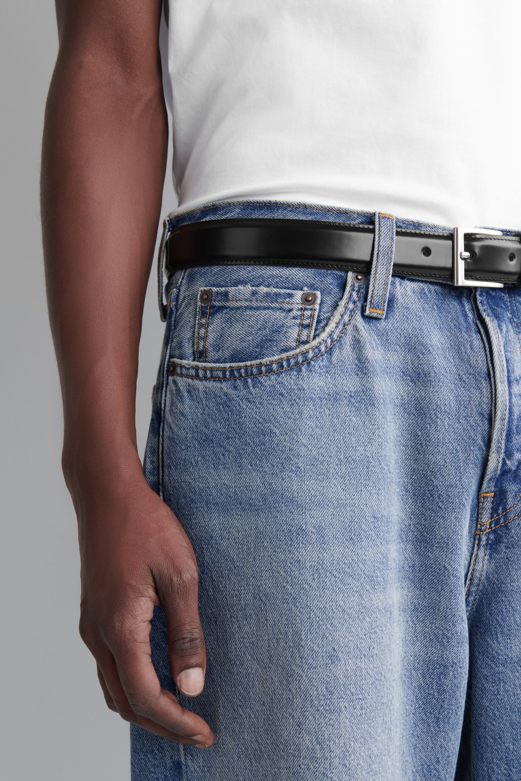 Visa större bild: Ljusblå denimjeans bärs, med en ljus tvätt, synliga framfickor med kontrasterande sömmar och nitar, och en avslappnad passform. Ett svart bälte med ett rektangulärt spänne i silverton träs genom bältesöglorna, och en vit t-shirt stoppas in i jeansen. Jeansen ses från höger framhöft, vilket framhäver deras avslappnade passform.