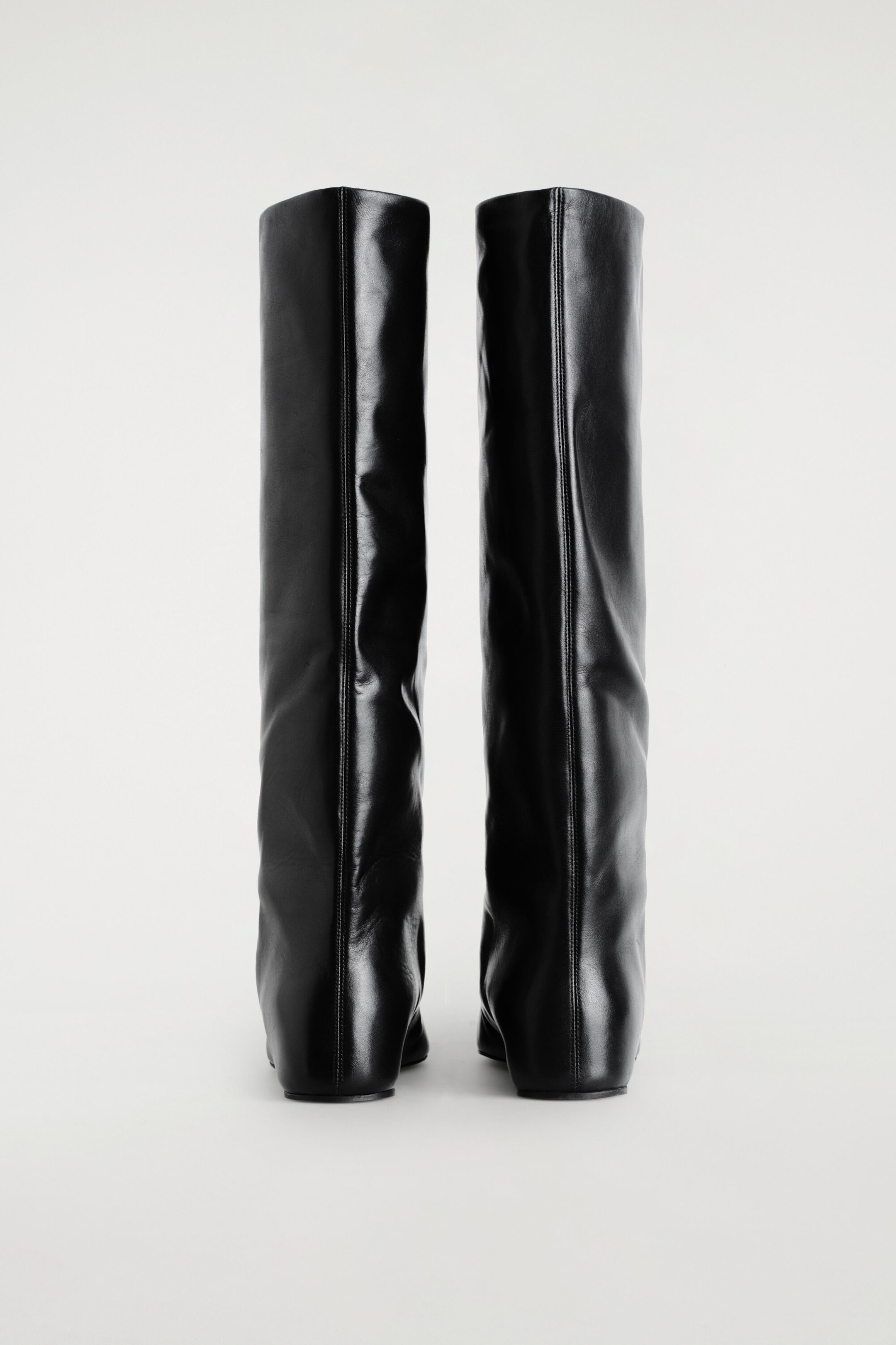 A pair of black leather knee-high boots stands upright, facing away from the viewer, showcasing a prominent vertical seam running down the center of each shaft. The boots feature a smooth finish with subtle wrinkling and low, covered wedge heels. A pair of black leather knee-high boots stands upright, facing away from the viewer, showcasing a prominent vertical seam running down the center of each shaft. The boots feature a smooth finish with subtle wrinkling and low, covered wedge heels.