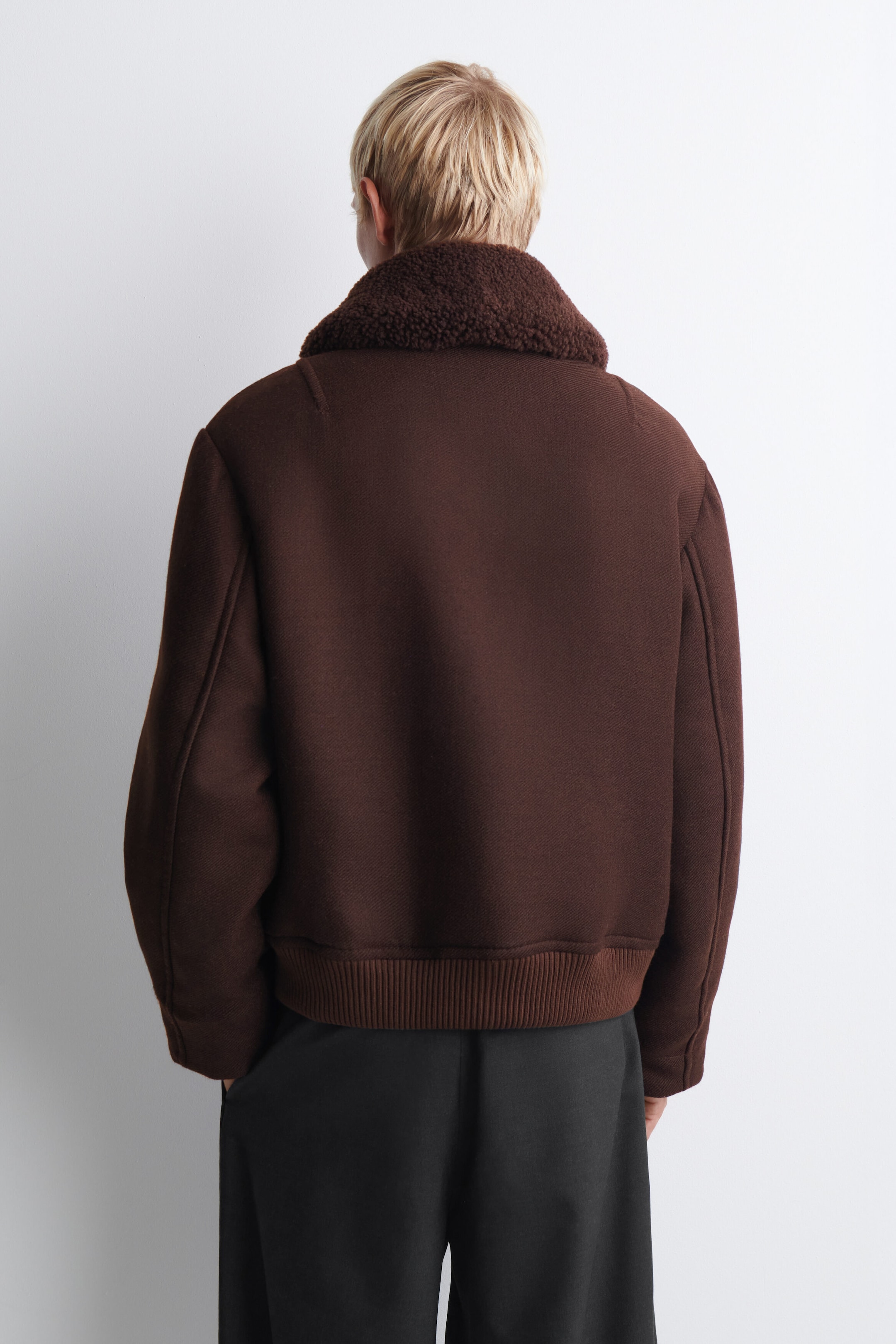 A dark brown bomber jacket is shown from the back, featuring a plush, textured sherpa collar and a wide ribbed knit hem. The jacket has a relaxed fit with structured seam detailing along the shoulders and sleeves. A dark brown bomber jacket is shown from the back, featuring a plush, textured sherpa collar and a wide ribbed knit hem. The jacket has a relaxed fit with structured seam detailing along the shoulders and sleeves.