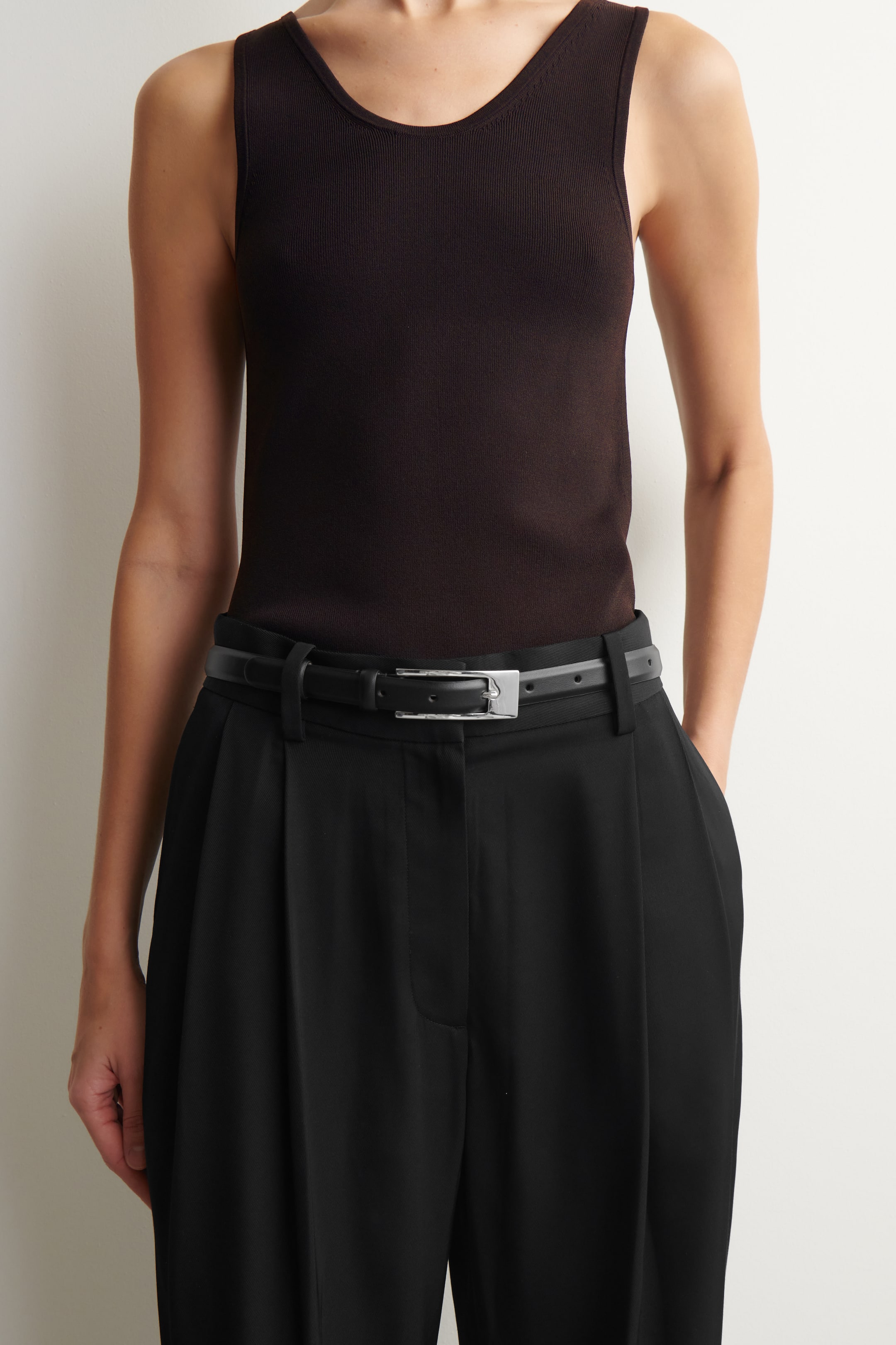 Agrandir l'image: Une ceinture en cuir noir est portée par le mannequin autour de sa taille, maintenant en place un pantalon plissé noir. Elle se caractérise par une sangle étroite et une boucle allongée de couleur argentée.