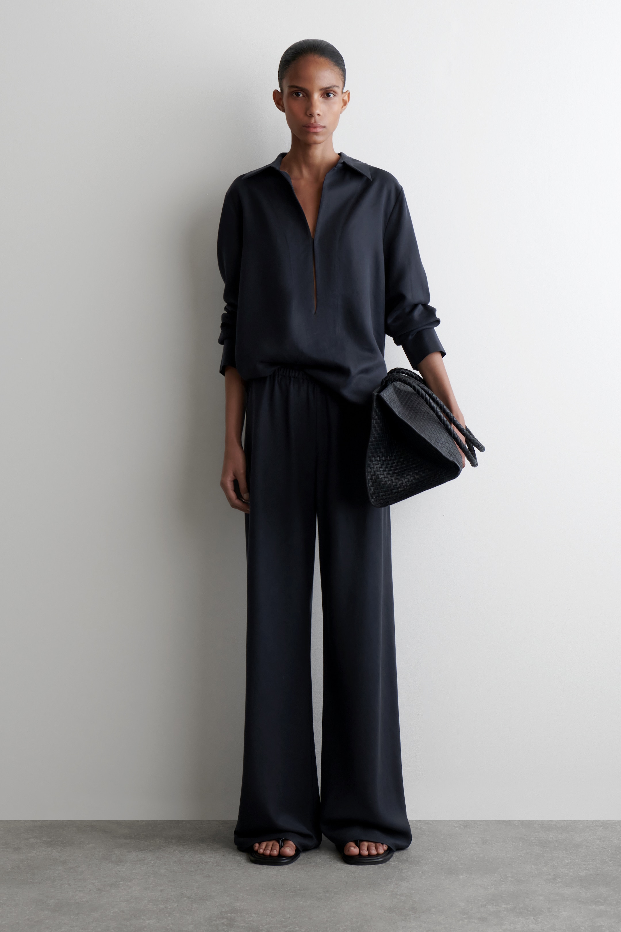 Agrandir l'image: Un ensemble deux pièces sombre et ample est présenté de face, comprenant un haut à manches longues avec un col évasé et un décolleté en V profond, ainsi qu'un pantalon large assorti avec une taille élastiquée. Un sac à main tissé sombre avec des poignées tressées est tenu, et des sandales noires à lanières sont portées avec la tenue.