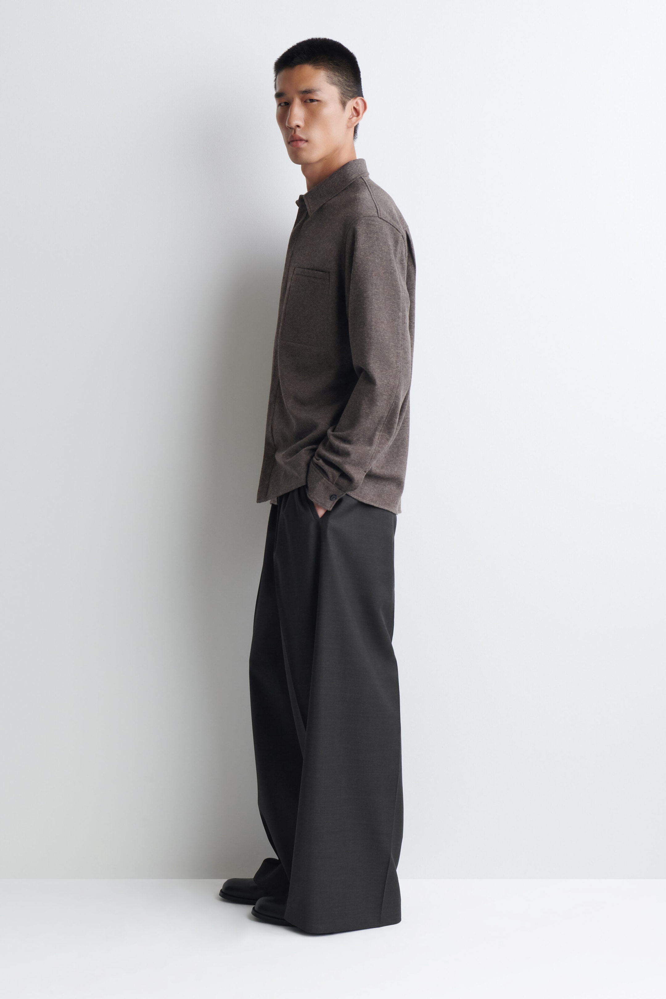 A brown long-sleeved shirt features a pointed collar, buttoned cuffs, and a left chest patch pocket. It is worn with black wide-leg trousers, which have a full-length silhouette that pools over the shoes. A brown long-sleeved shirt features a pointed collar, buttoned cuffs, and a left chest patch pocket. It is worn with black wide-leg trousers, which have a full-length silhouette that pools over the shoes.
