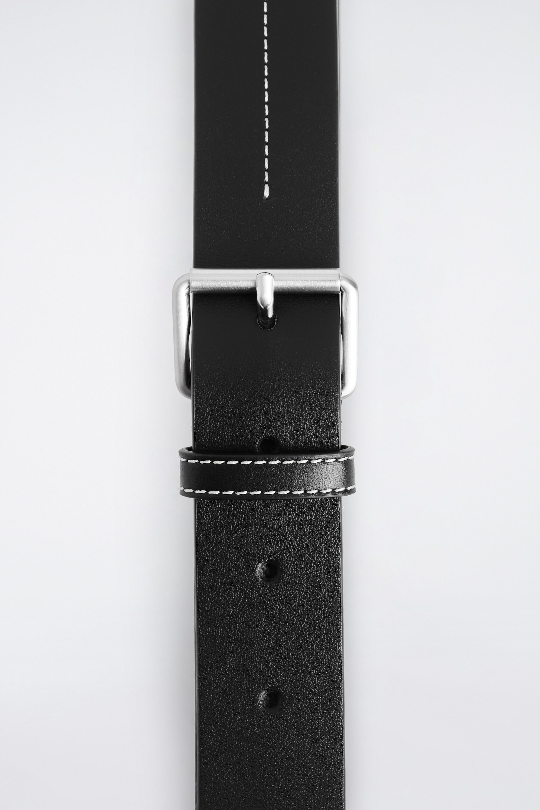 A black leather belt extends vertically, featuring a silver-toned metal buckle and a matching leather keeper with white contrast stitching. The upper strap shows a vertical line of white contrast stitching down the centre, while the lower strap shows several adjustment holes. A black leather belt extends vertically, featuring a silver-toned metal buckle and a matching leather keeper with white contrast stitching. The upper strap shows a vertical line of white contrast stitching down the centre, while the lower strap shows several adjustment holes.