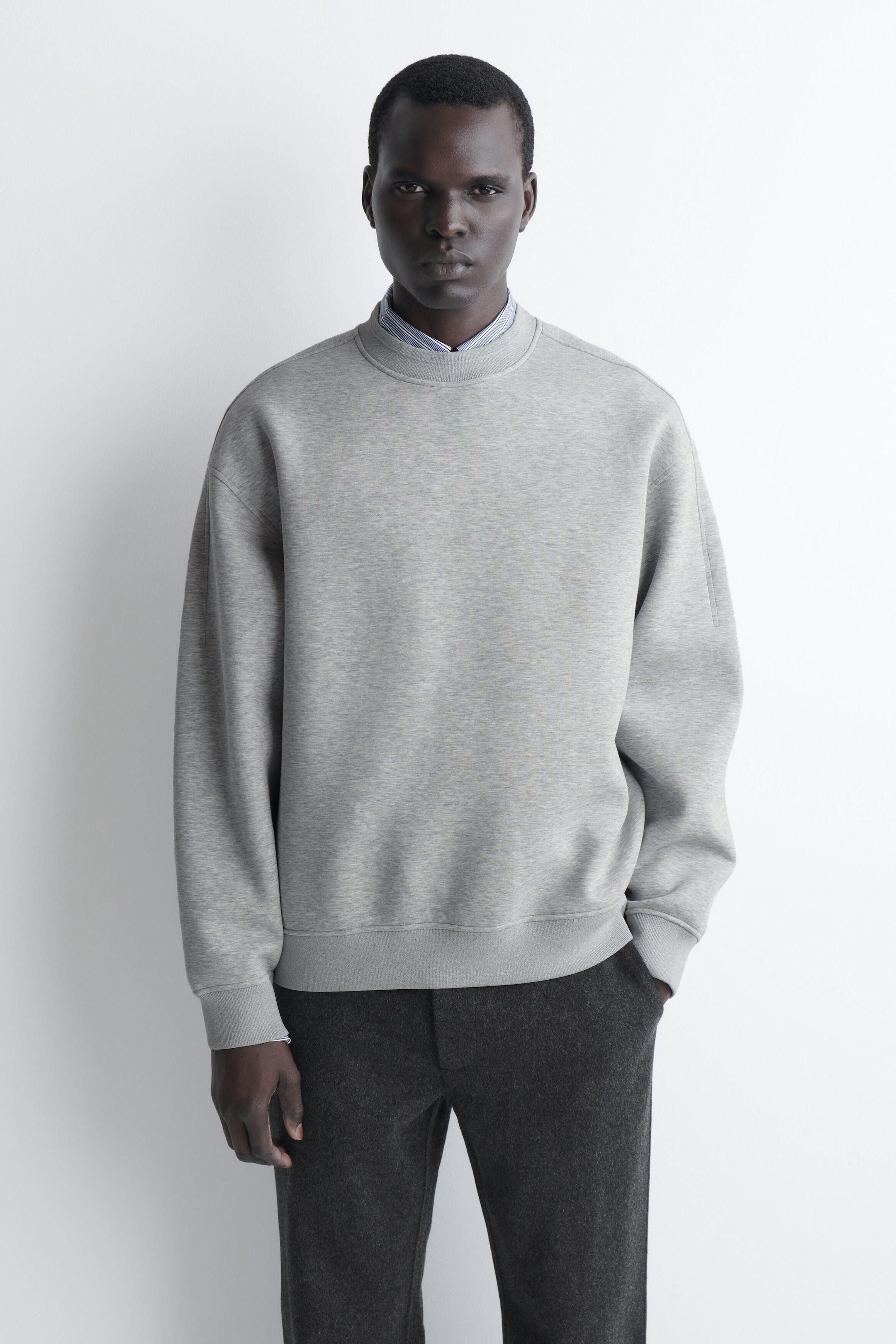 The relaxed scuba sweatshirt in light grey is shown in a loose fit with dropped shoulders, ribbed cuffs, and a ribbed hem from the front. A striped shirt collar peeks out from underneath the crewneck, illustrating a layered style. The relaxed scuba sweatshirt in light grey is shown in a loose fit with dropped shoulders, ribbed cuffs, and a ribbed hem from the front. A striped shirt collar peeks out from underneath the crewneck, illustrating a layered style.