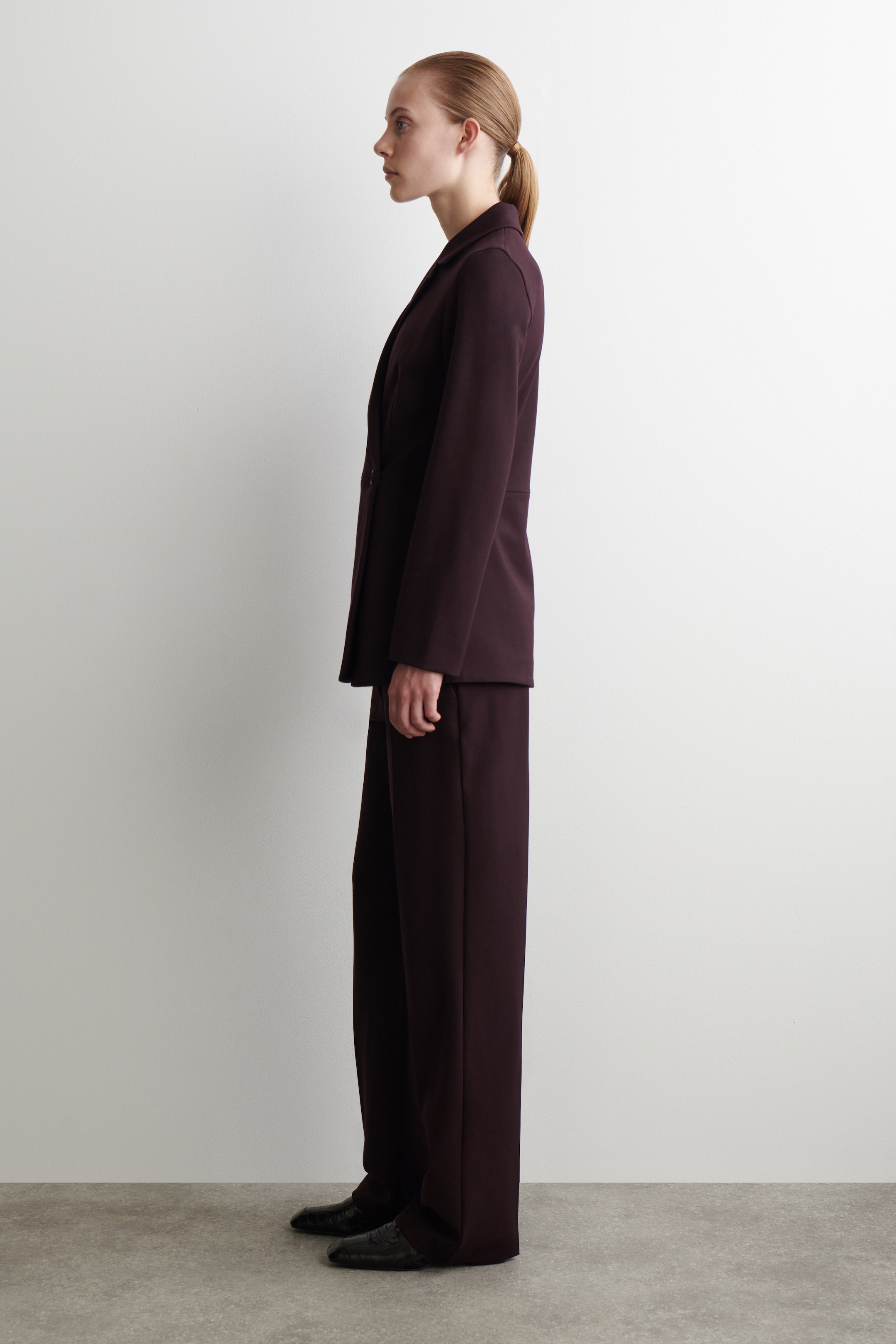 Visualizza immagine più grande: Il blazer color bordeaux viene presentato dal lato sinistro, caratterizzato da un tessuto fluido, revers a dente e una linea di cintura definita, ed è abbinato come un completo con pantaloni a gamba larga coordinati.