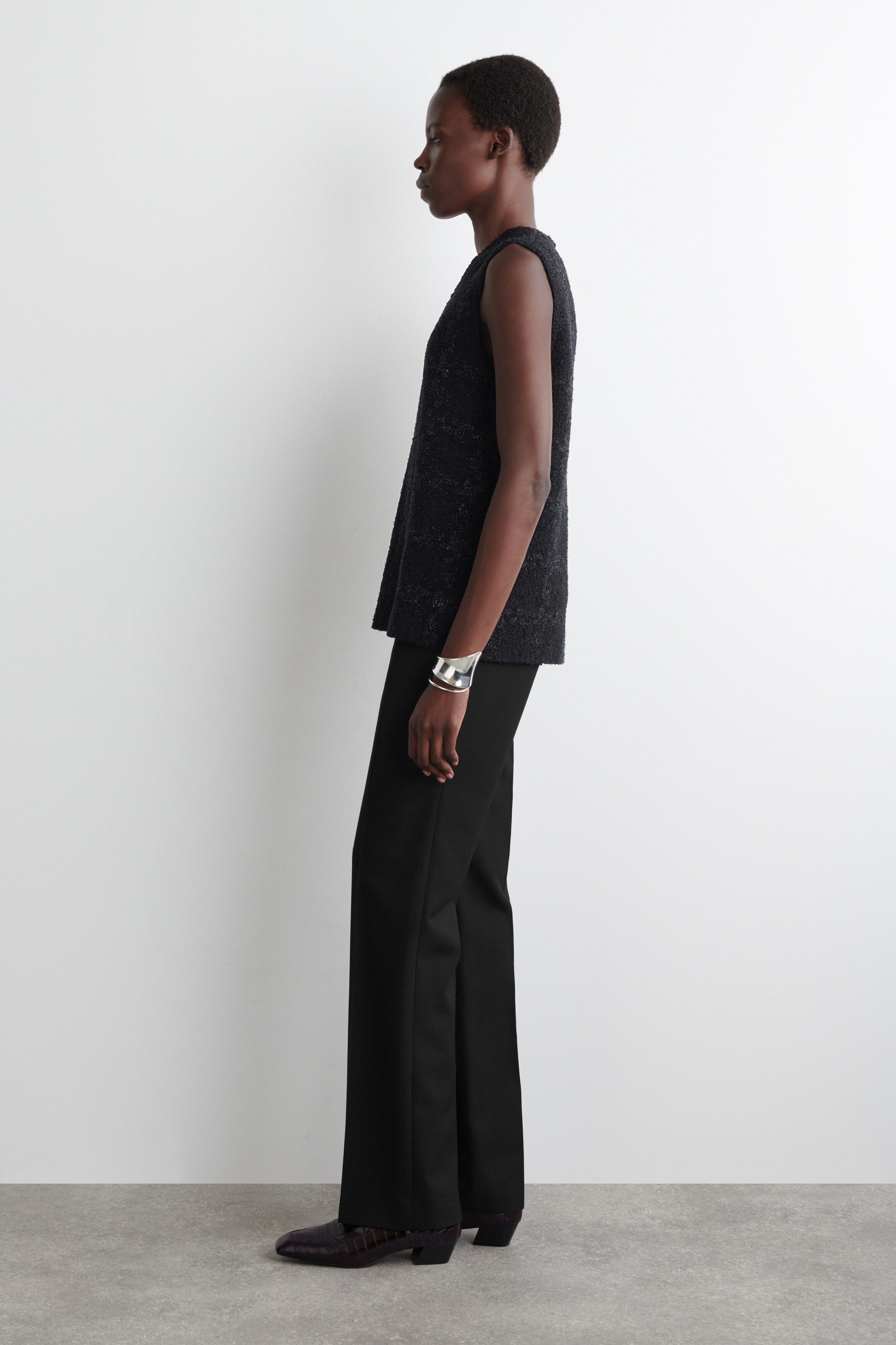 Agrandir l'image: Un haut sans manches sombre et texturé est présenté de profil, stylisé avec un pantalon noir à jambes larges. Un large bracelet-manchette en argent est porté au poignet droit, et des chaussures à motifs sombres complètent l'ensemble.