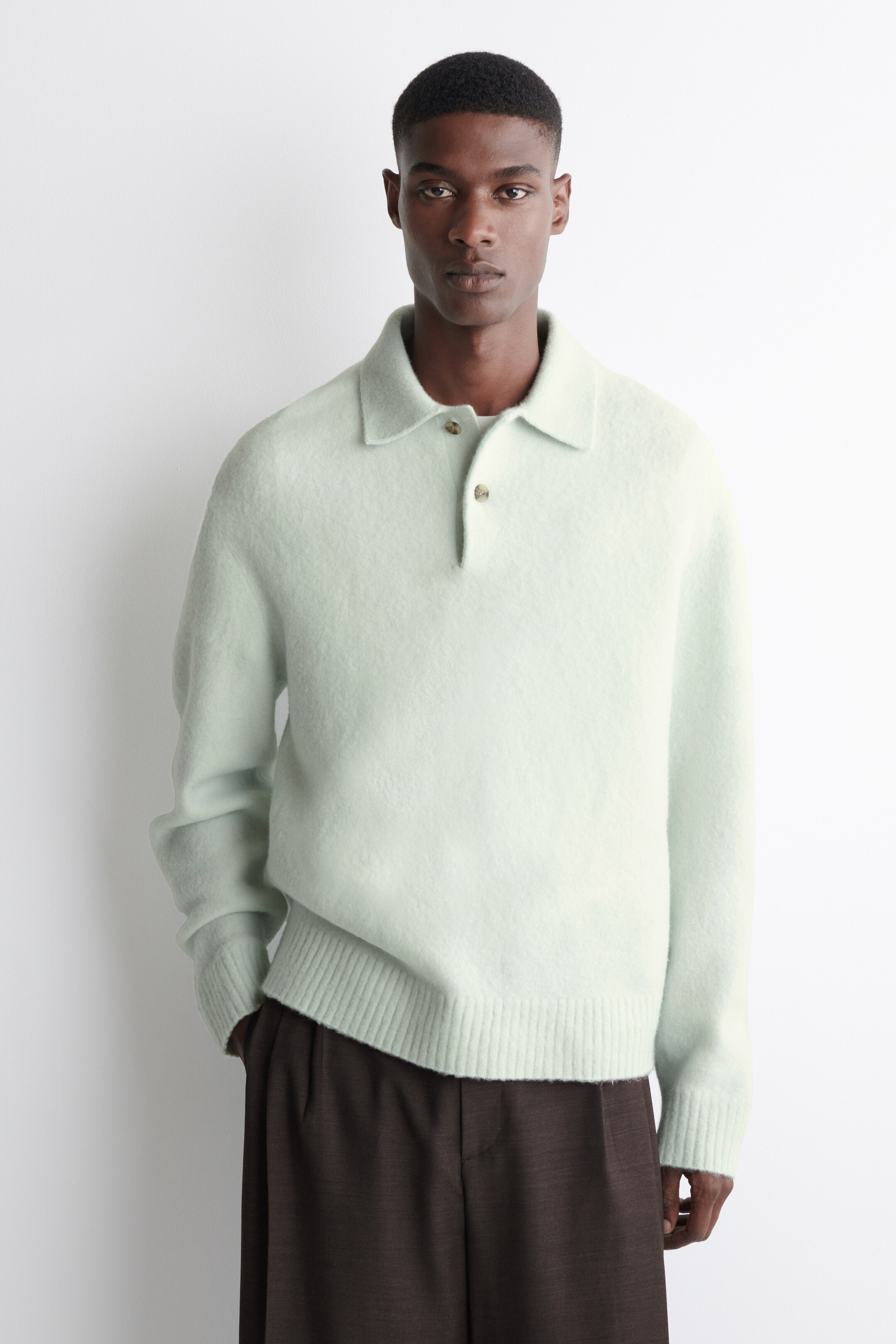 Visualizza immagine più grande: Un maglione polo verde chiaro viene indossato, rivolto in avanti. Presenta una texture lavorata a maglia pelosa, un classico colletto polo, un carré a due bottoni con bottoni marrone chiaro e polsini e orlo a coste.