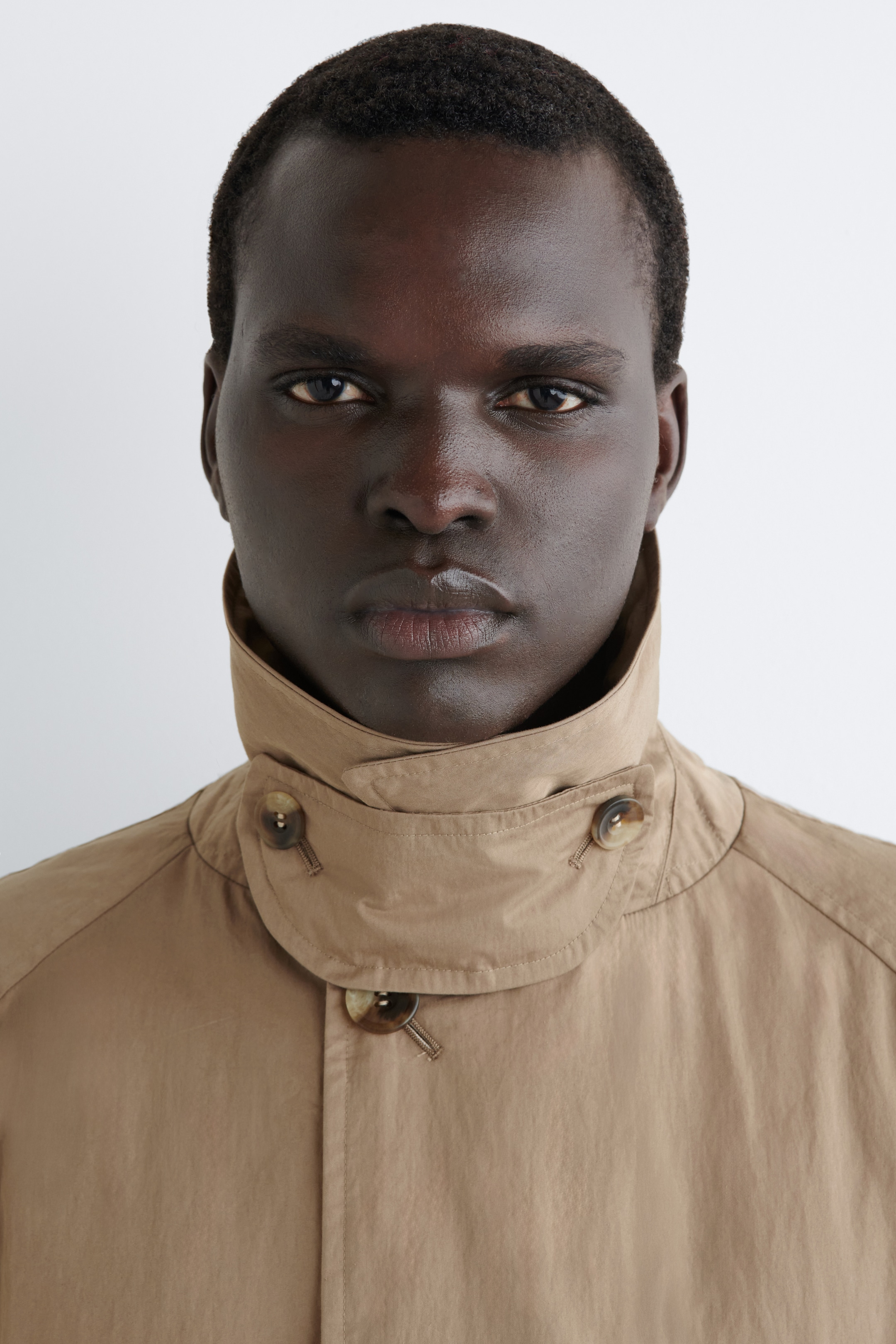 Visualizza immagine più grande: Un cappotto beige presenta un colletto alto e in piedi con una chiusura per la gola fissata da un bottone marrone marmorizzato. Un altro bottone coordinato è visibile sotto sulla liscia patta anteriore del cappotto.
