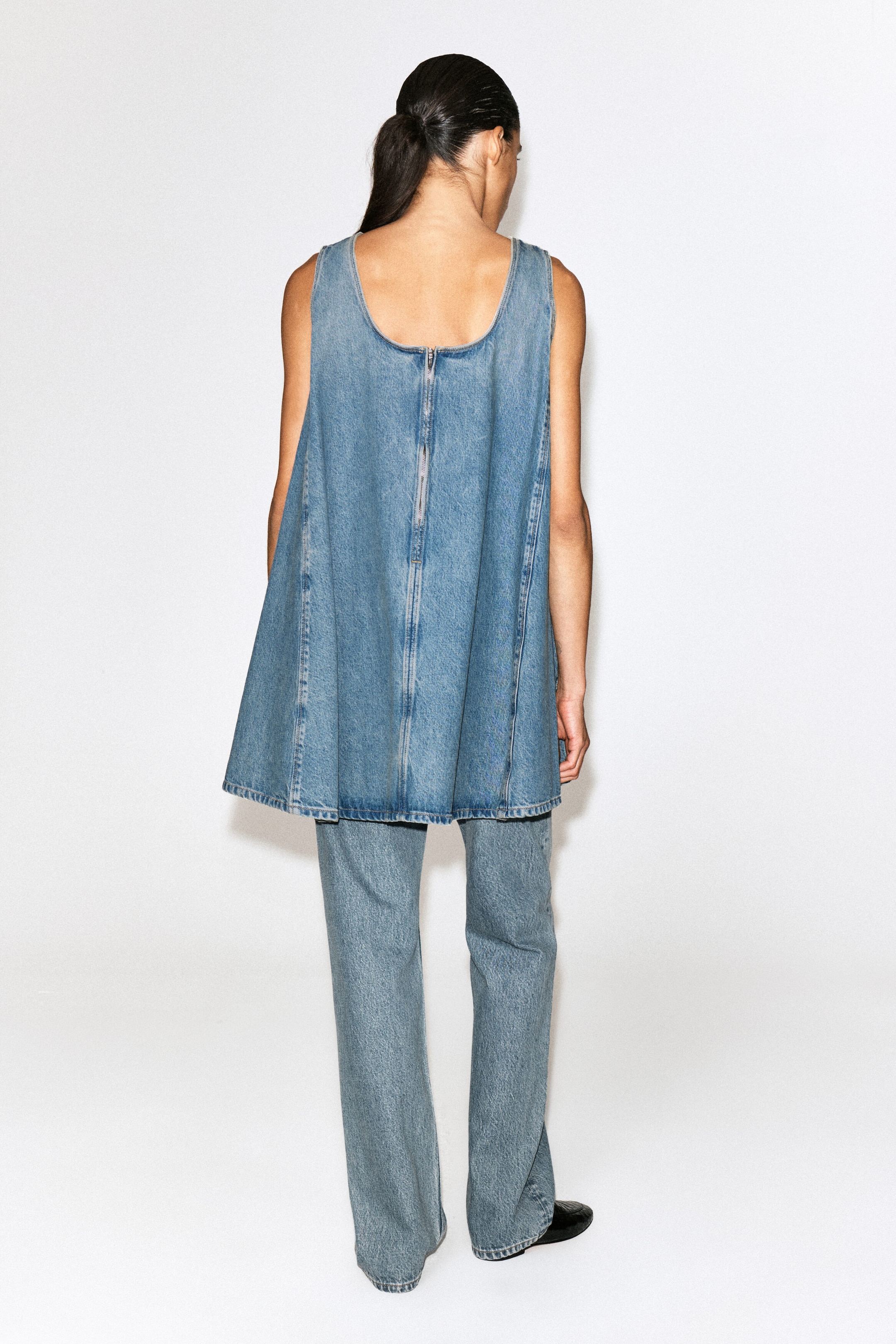 Un mini abito in denim senza maniche nel lavaggio azzurro chiaro è mostrato da dietro, con profonda scollatura sulla schiena, zip centrale a vista e cuciture verticali che si aprono in una silhouette a trapezio. L’abito è abbinato a jeans in denim coordinati a gamba dritta.