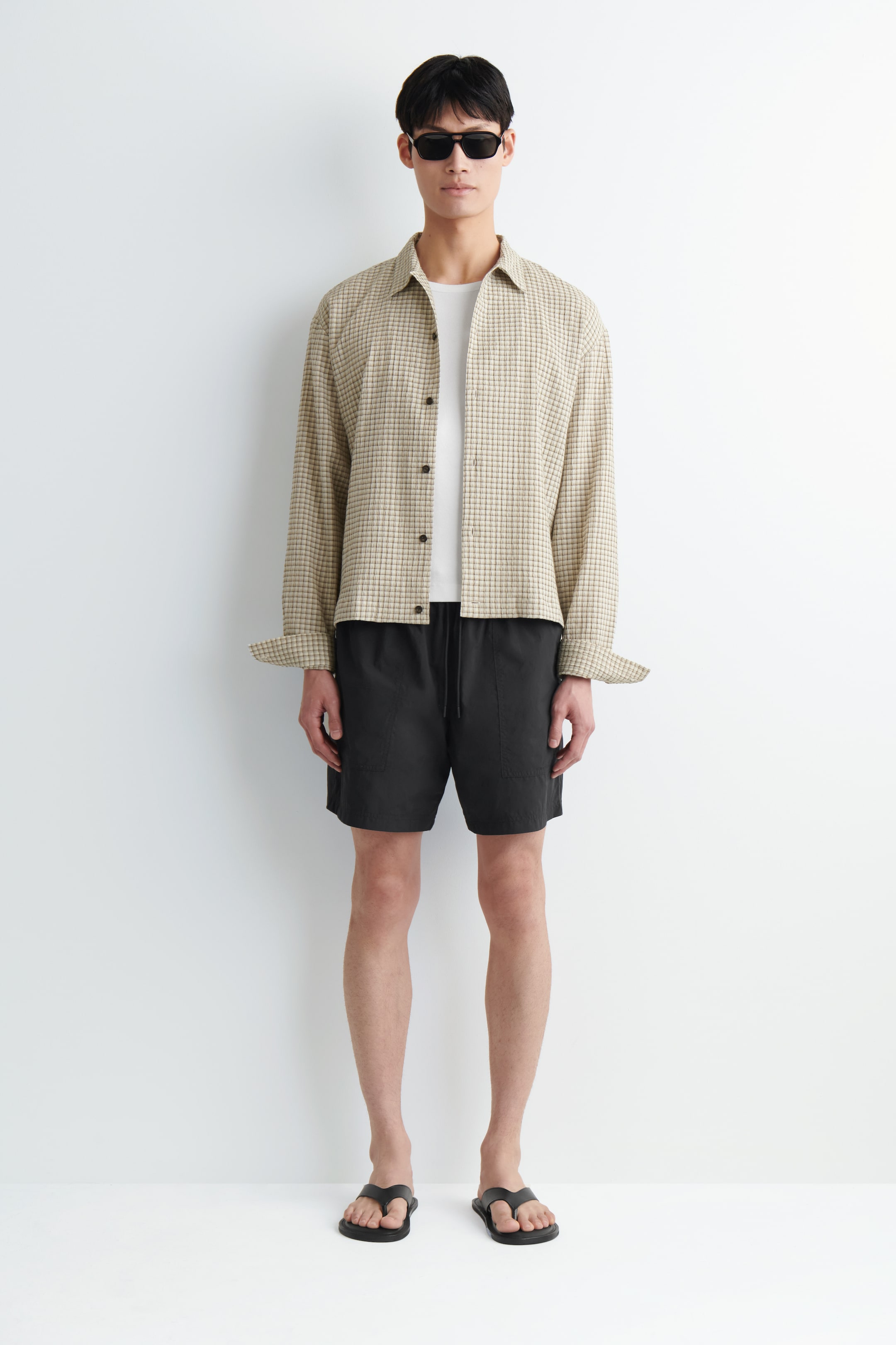 Visualizza immagine più grande: Una camicia a maniche lunghe a micro quadretti marrone chiaro e bianca è indossata aperta sopra una t-shirt bianca a girocollo, con i polsini arrotolati. Pantaloncini color carbone scuro con vita a coulisse e tasche frontali completano il look, abbinati a infradito piatte nere e occhiali da sole rettangolari.