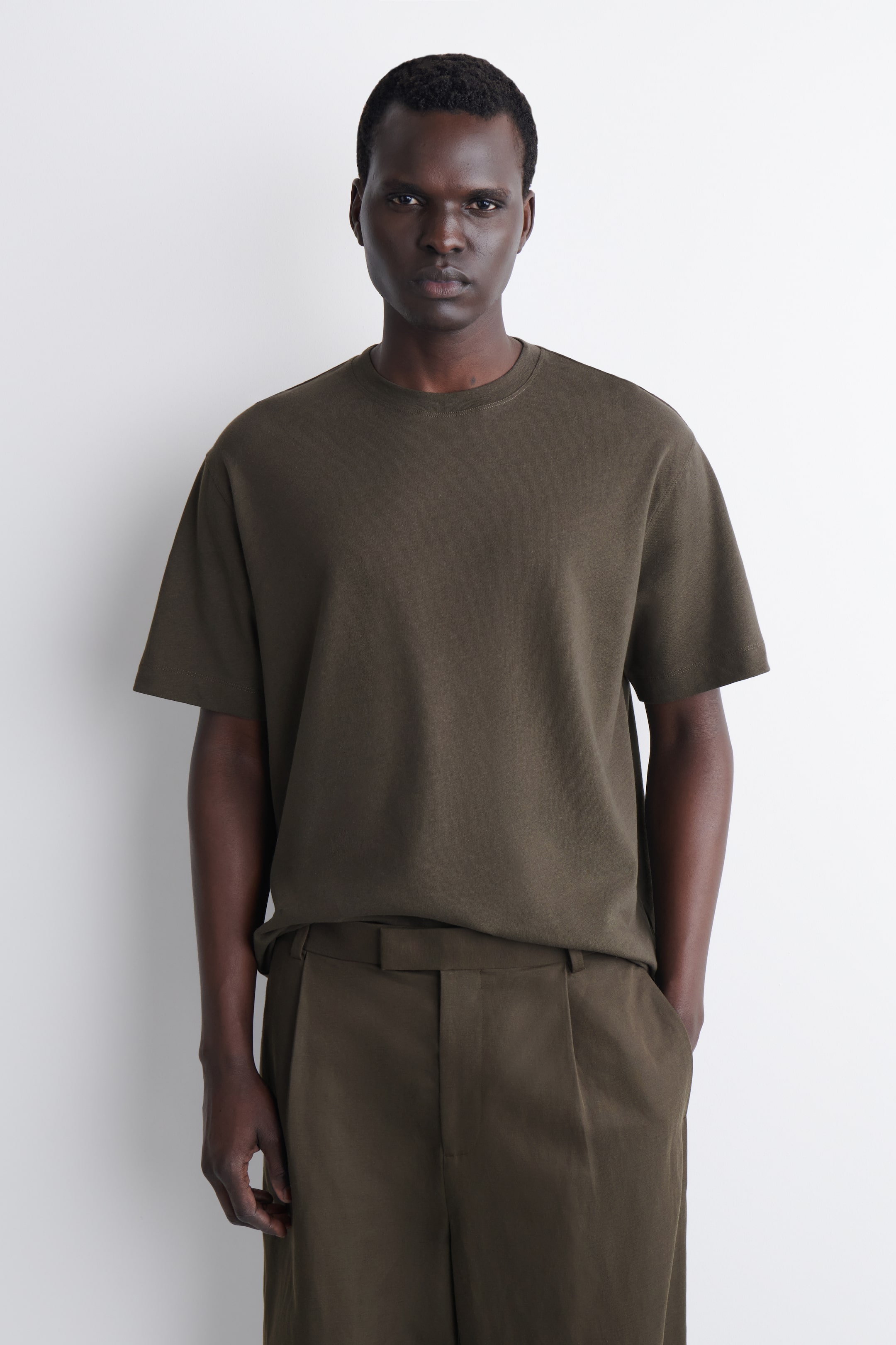 Größeres Bild ansehen: Ein dunkel olivgrünes T-Shirt mit kurzen Ärmeln, das einen Rundhalsausschnitt und eine entspannte Passform aufweist, wird von vorne präsentiert und mit passenden, plissierten Hosen gestylt.