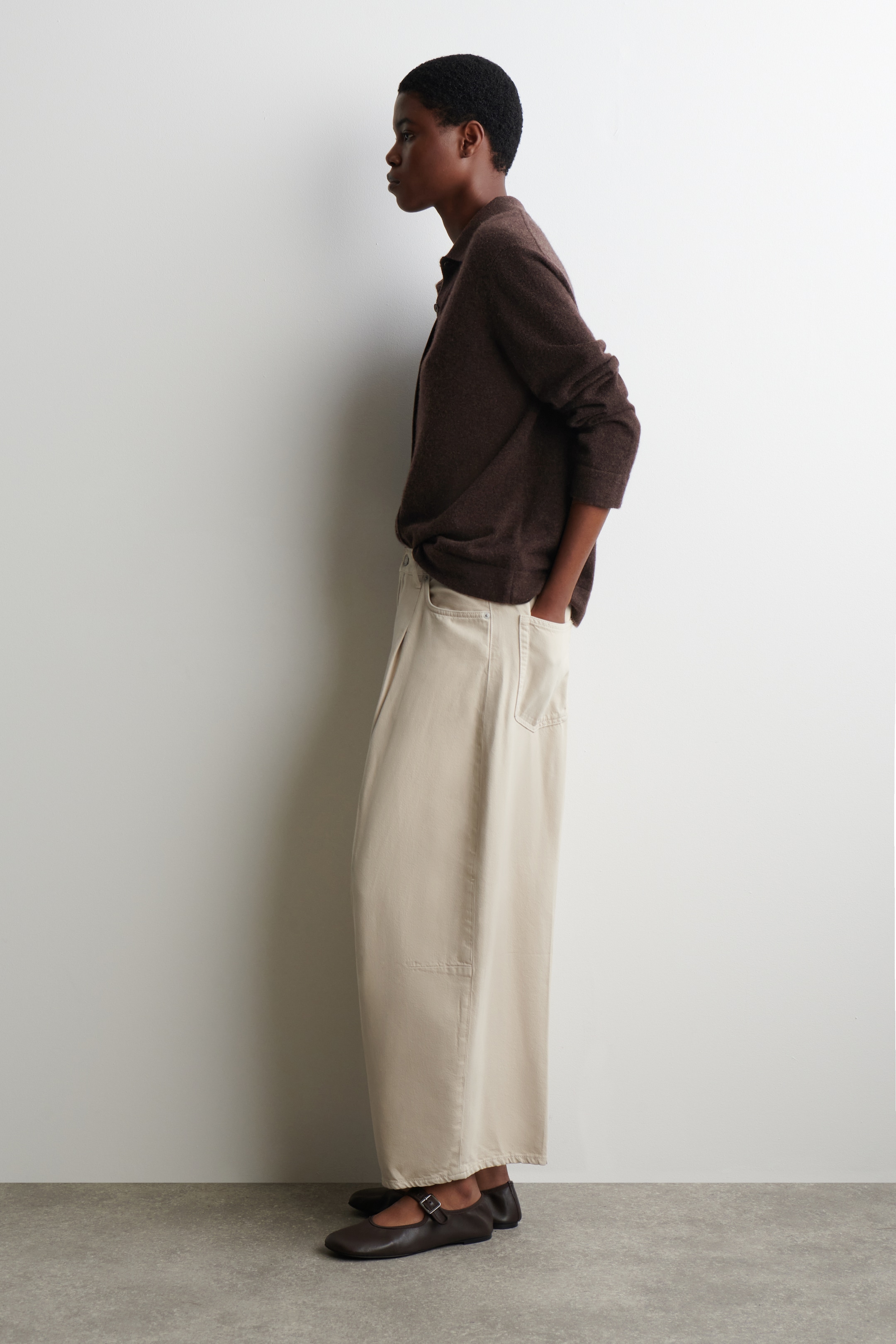 View larger image: A dark brown, long-sleeved collared shirt is being worn loosely and tucked into light beige, wide-leg pants featuring front pleats and side pockets. The outfit, shown from a side profile, is completed with dark brown Mary Jane flat shoes that have a buckled strap.